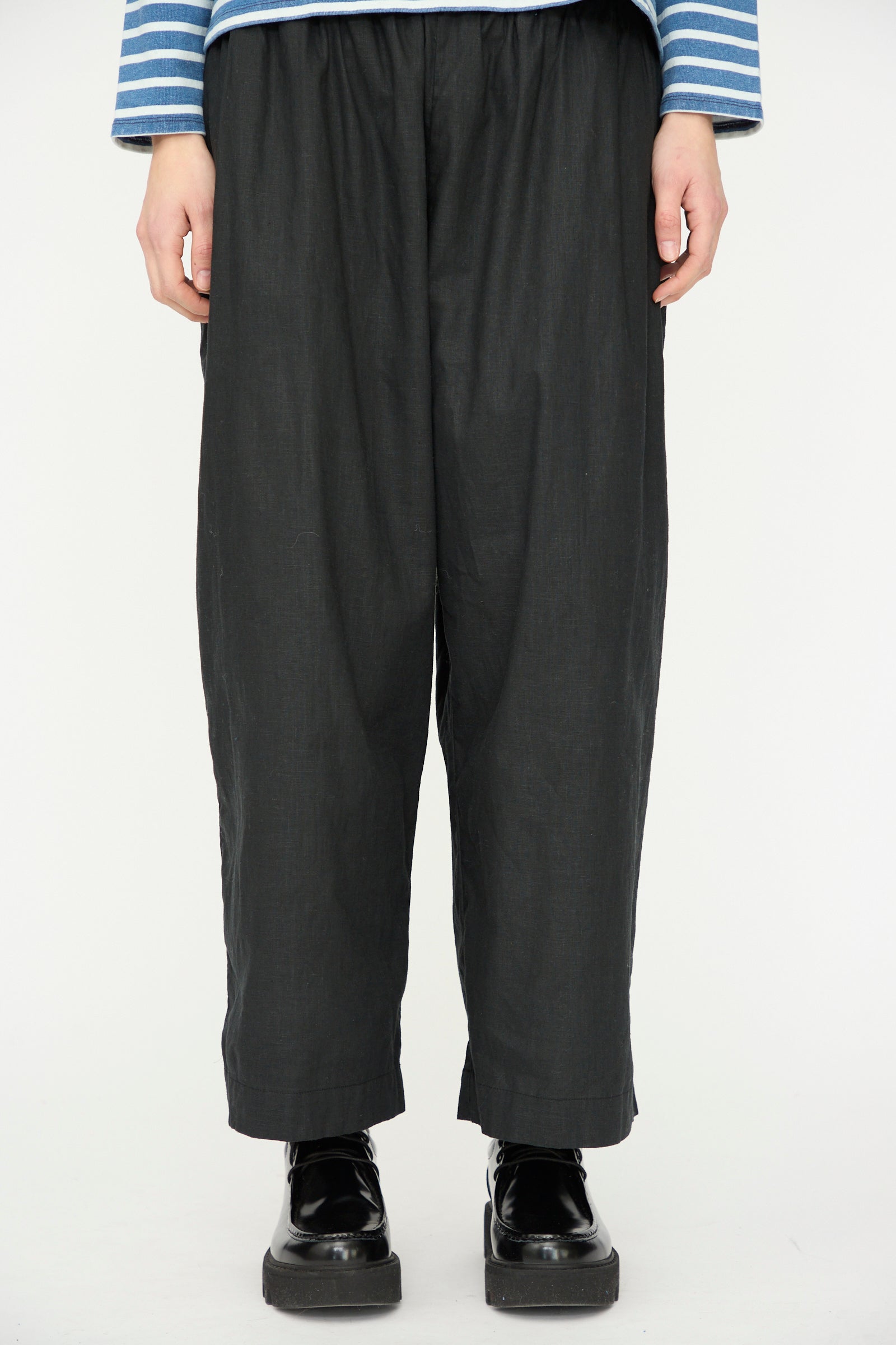 A person wearing the Ichi Antiquités Linen Cotton Canvas Pant in Black, paired with a blue and white striped long-sleeve shirt and black platform shoes, stands against a plain white background.