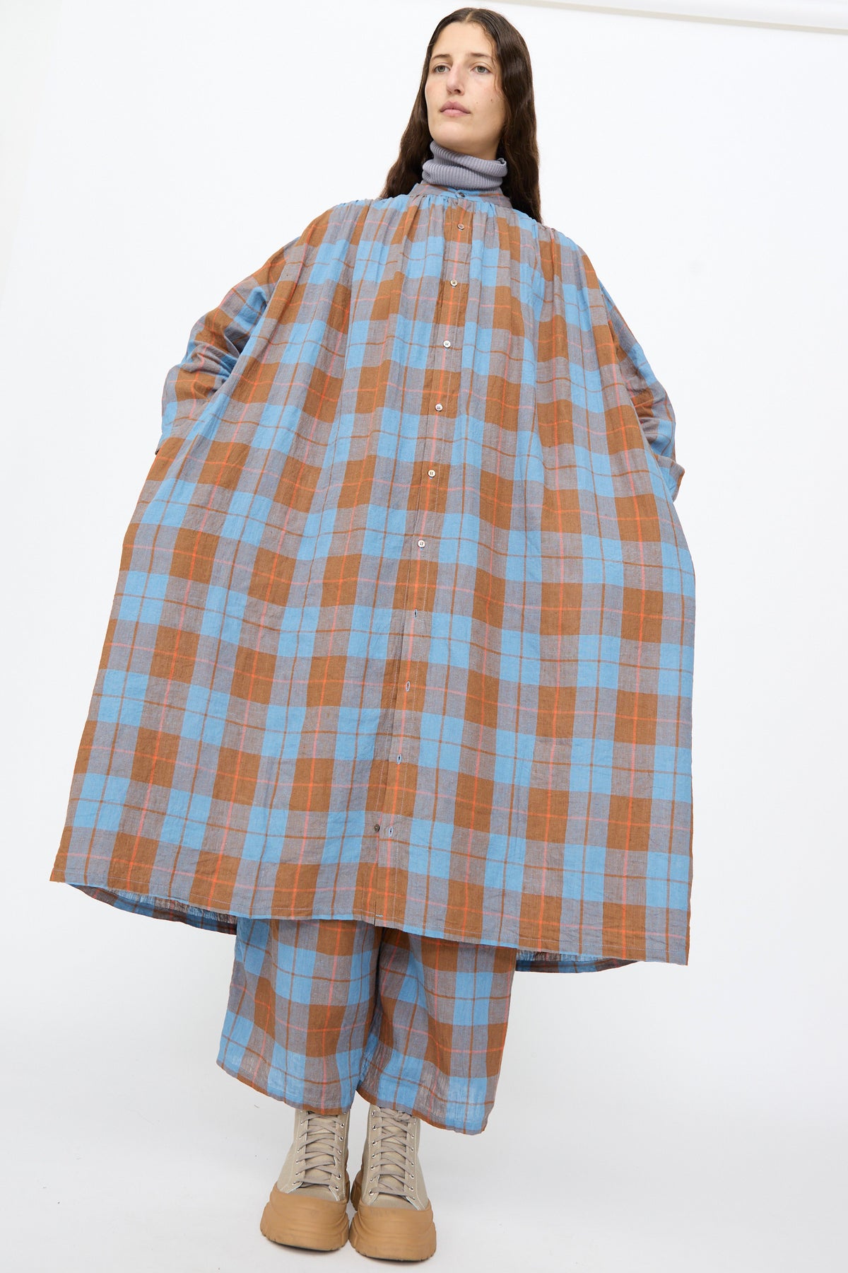 A person wears an oversized, long-sleeve Ichi Antiquités Linen Cotton Check Dress in blue and brown plaid over matching trousers and beige high-top sneakers.