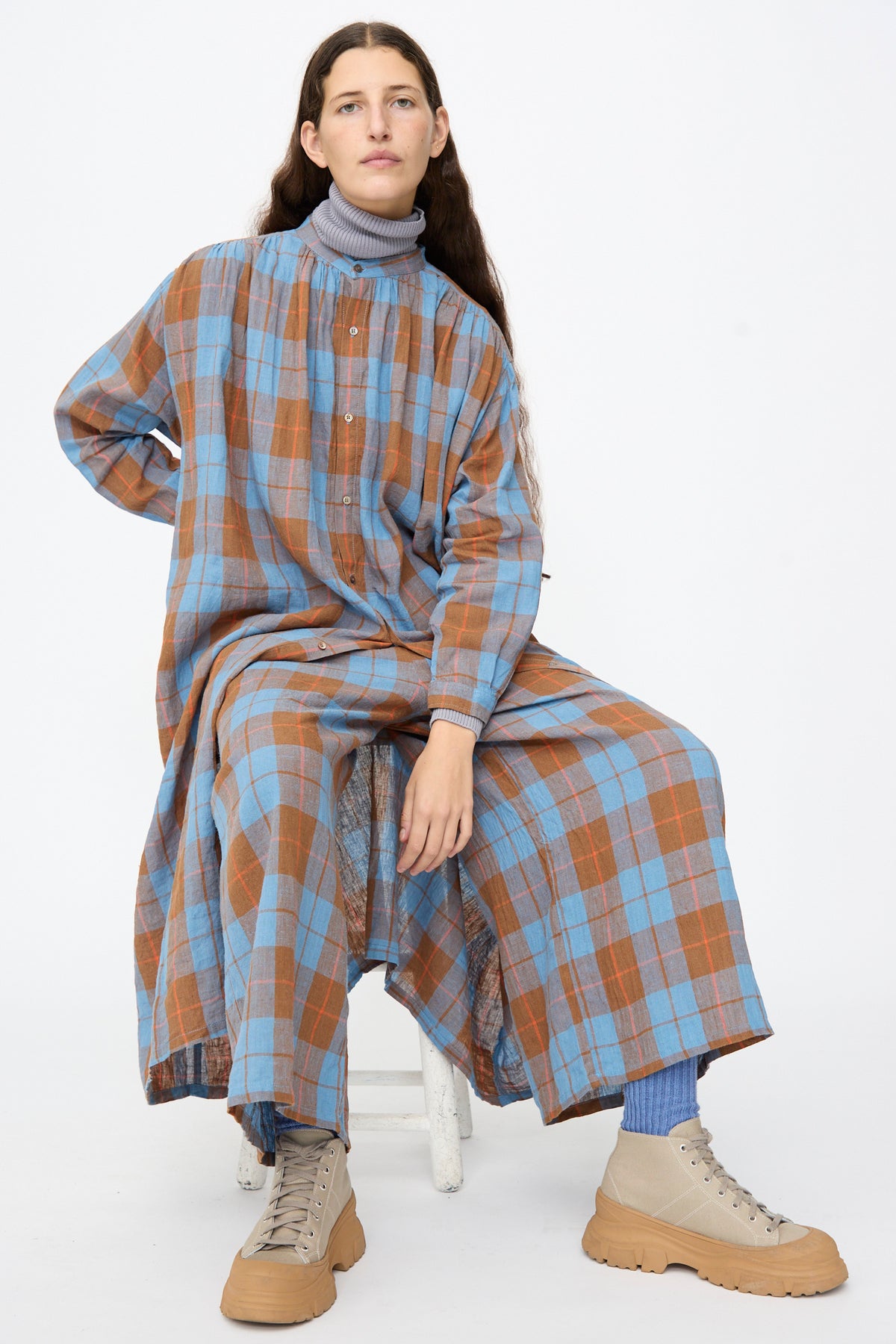 A person wearing an Ichi Antiquités Linen Cotton Check Dress in Blue over a grey mock-neck top, with blue socks and beige sneakers, sitting on a white stool.