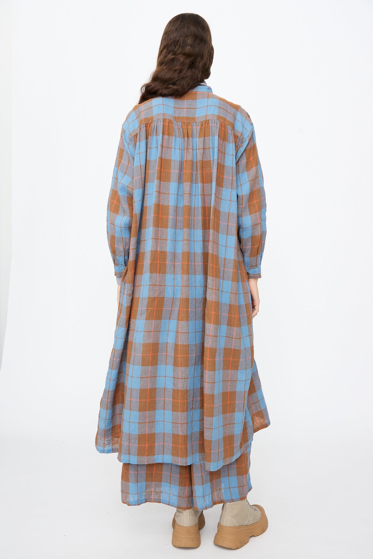 A person wearing an Ichi Antiquités Linen Cotton Check Dress in Blue, a long-sleeved, blue and brown plaid midi dress with a gathered back yoke, paired with tan platform boots.