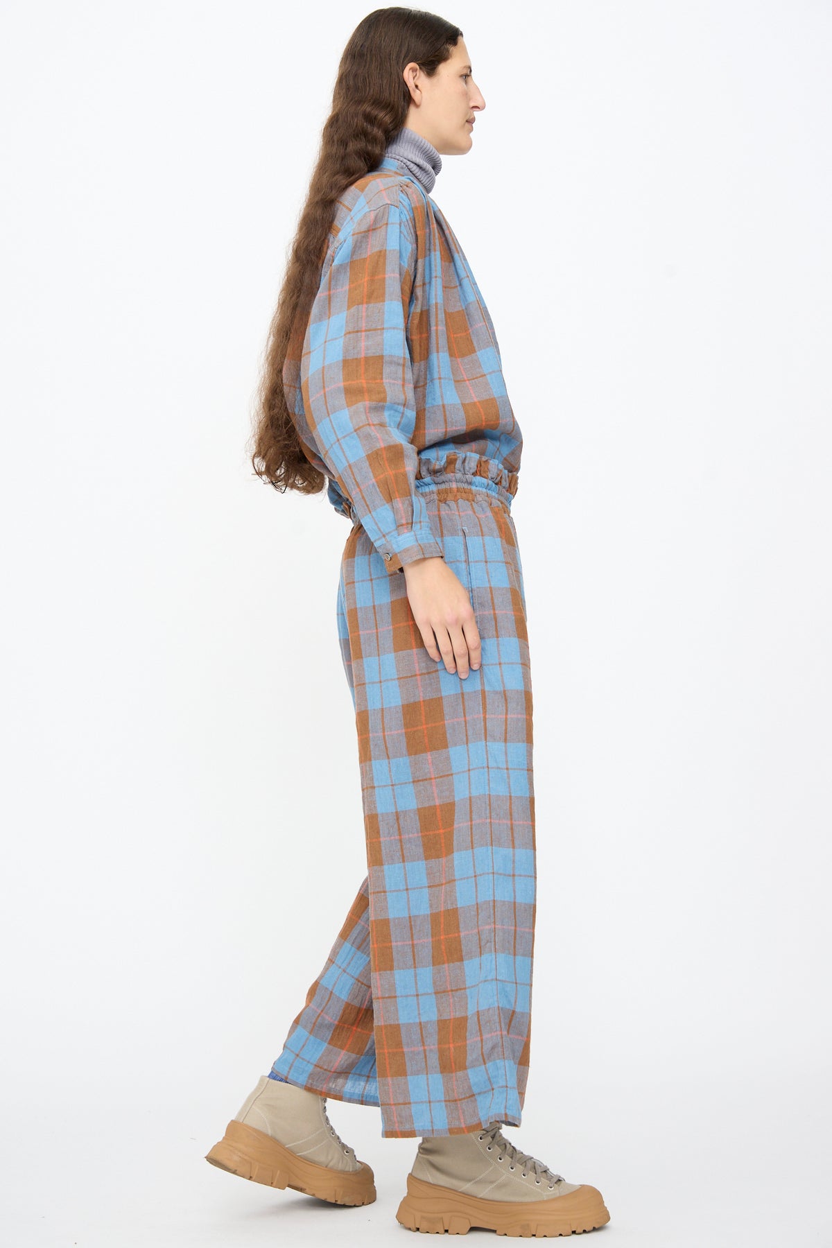 A person wearing a blue and brown plaid long-sleeved shirt and matching Ichi Antiquités Linen Cotton Check Pants in Blue, styled with a grey turtleneck and tan lace-up boots.