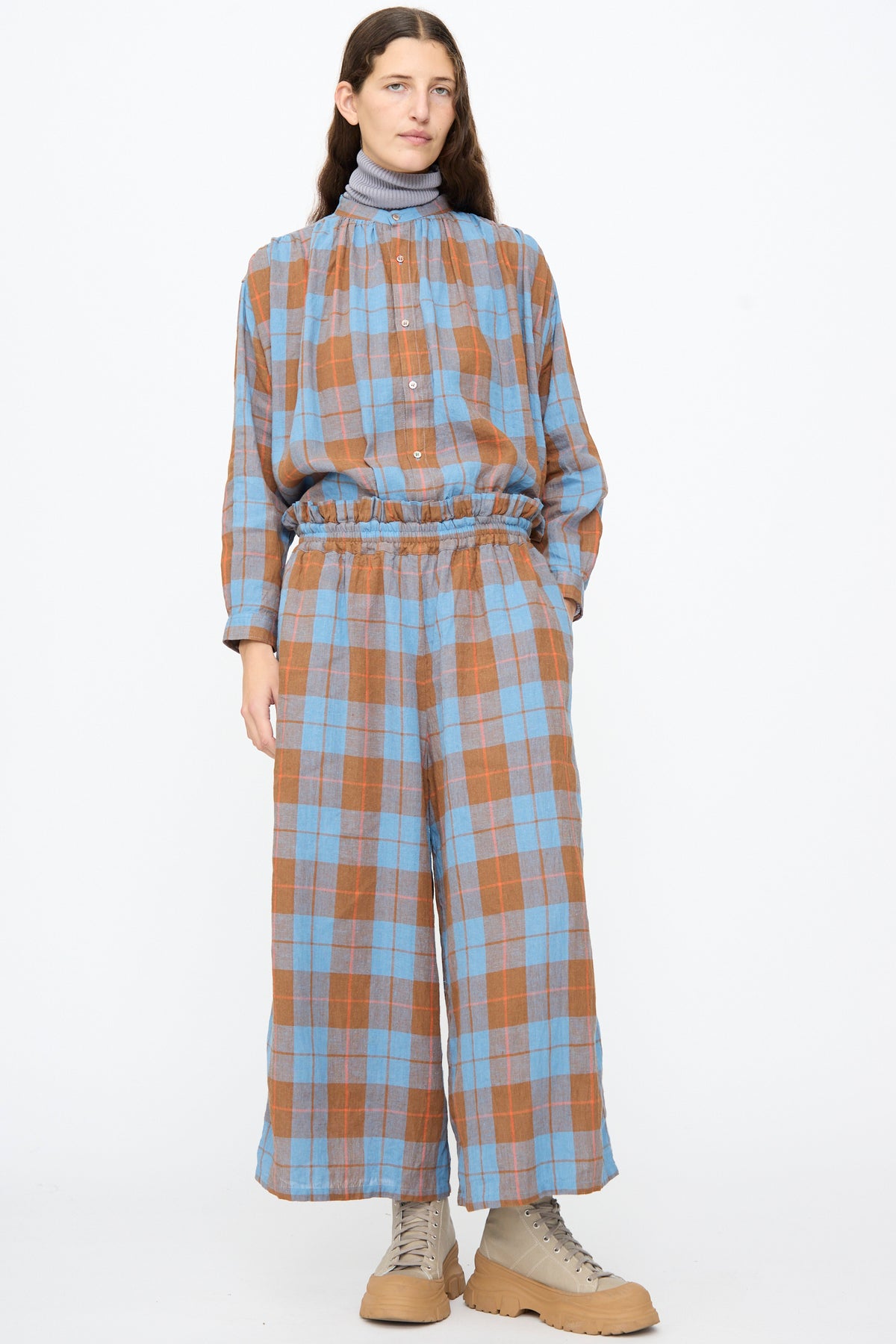 A person wears a matching long-sleeved button-up shirt and Ichi Antiquités Linen Cotton Check Pants in Blue, paired with a grey turtleneck and beige boots.