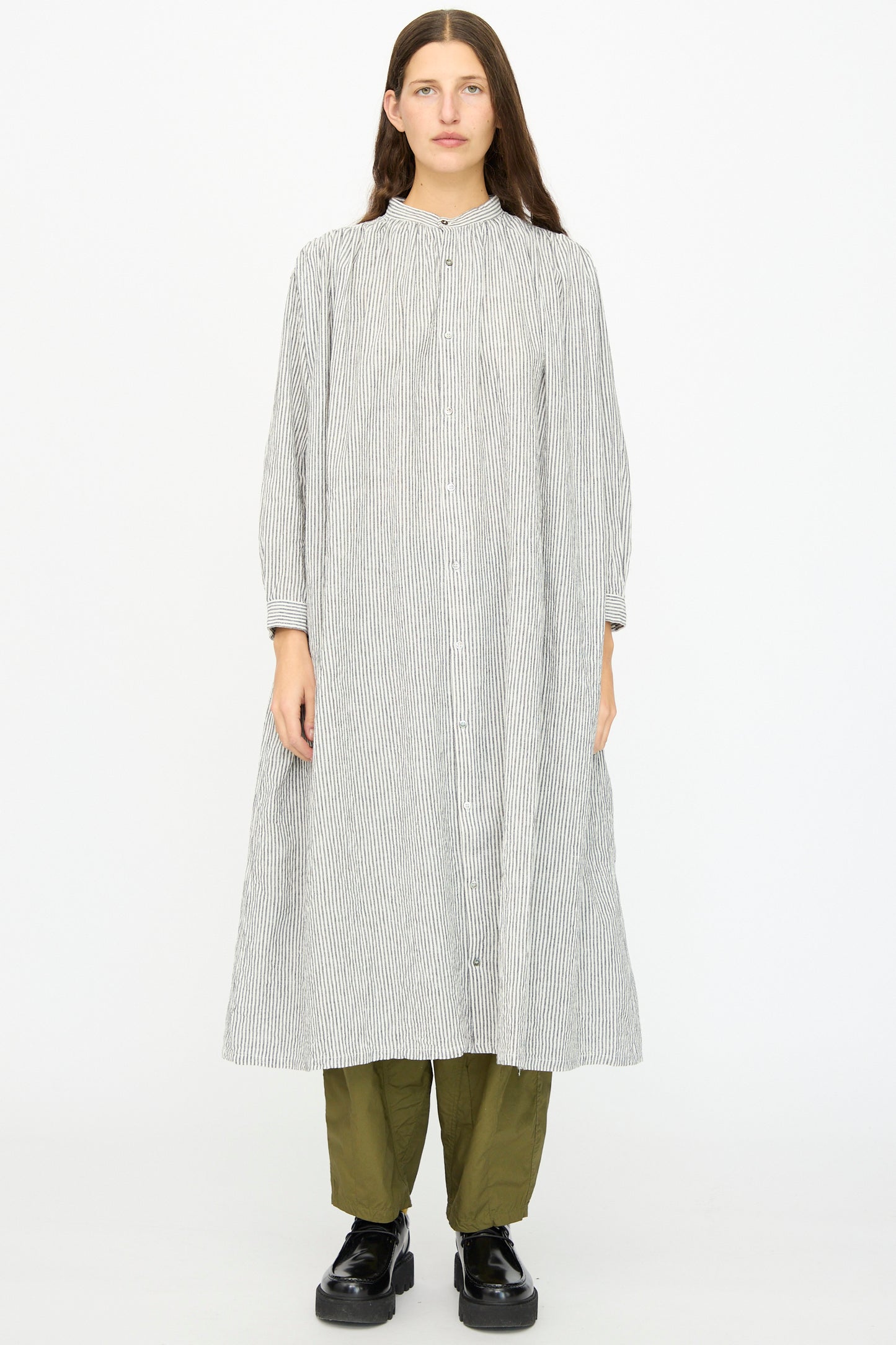 A person faces forward, wearing the Ichi Antiquités Linen and Cotton Dress in White Stripe, layered over olive green pants and black shoes, set against a plain white background.