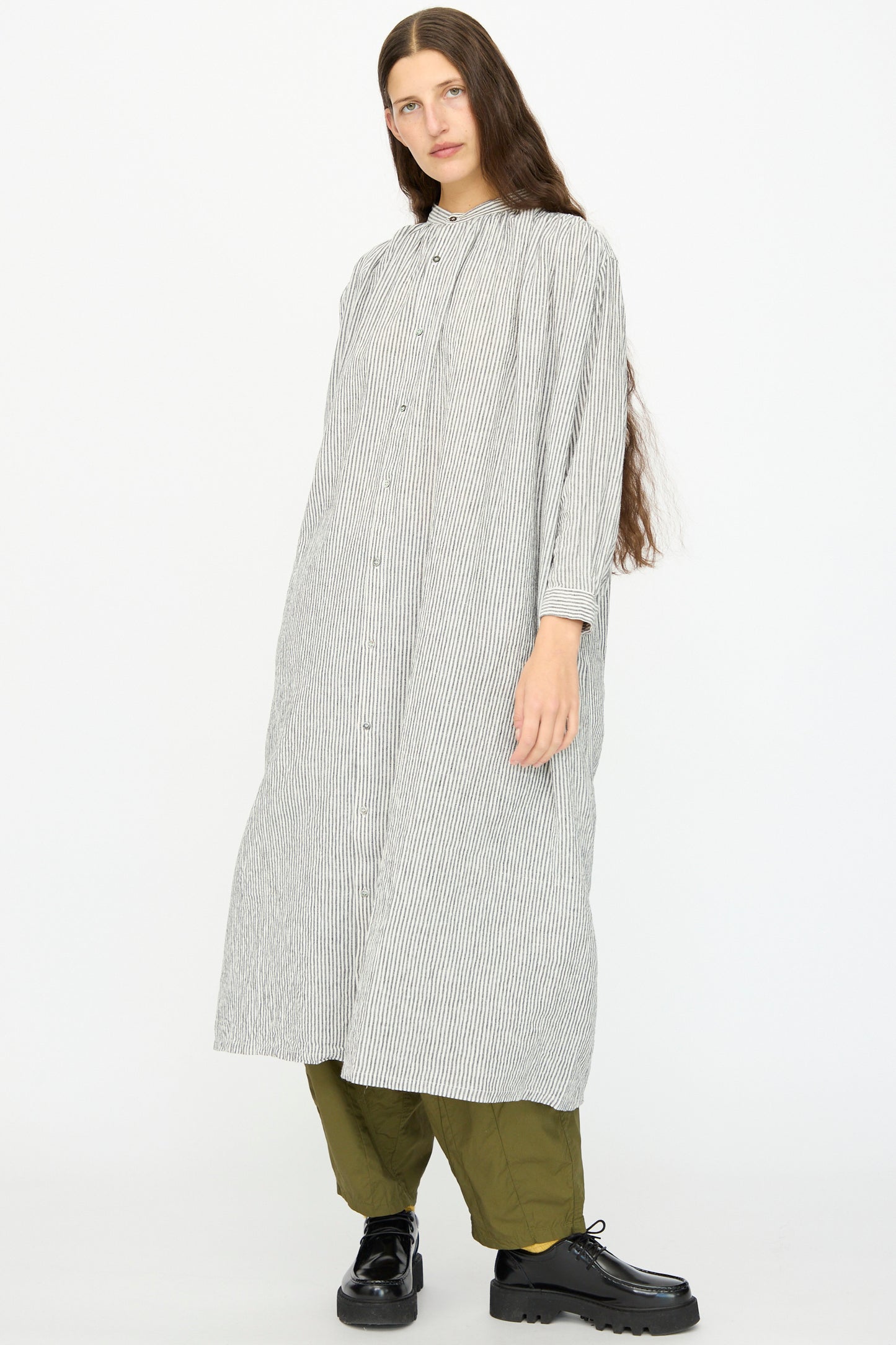 A person wears the Ichi Antiquités Linen and Cotton Dress in White Stripe over olive green pants and black shoes, standing against a plain white background.