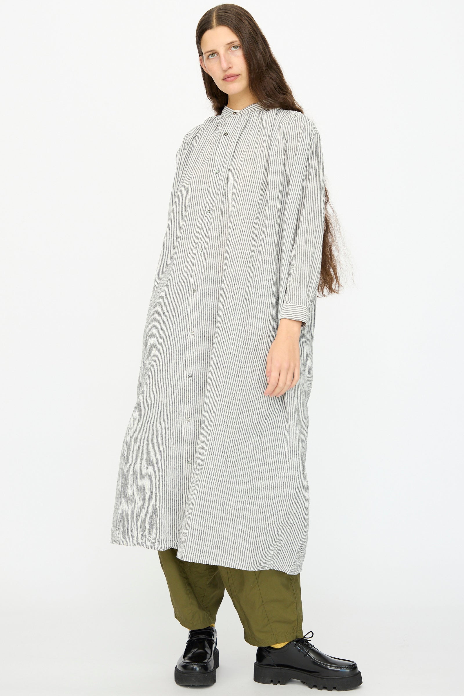 A person wears the Ichi Antiquités Linen and Cotton Dress in White Stripe over olive green pants and black shoes, standing against a plain white background.