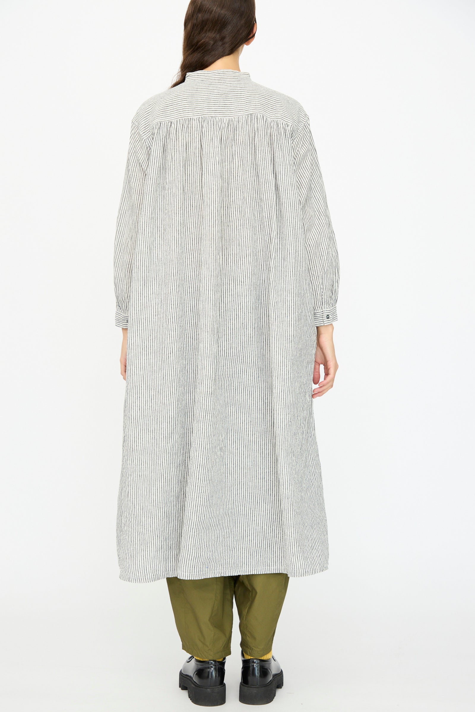 A person with tied-back brown hair stands facing away, wearing the Ichi Antiquités Linen and Cotton Dress in White Stripe over olive green pants and black shoes—crafted in Japan for effortless style and comfort.