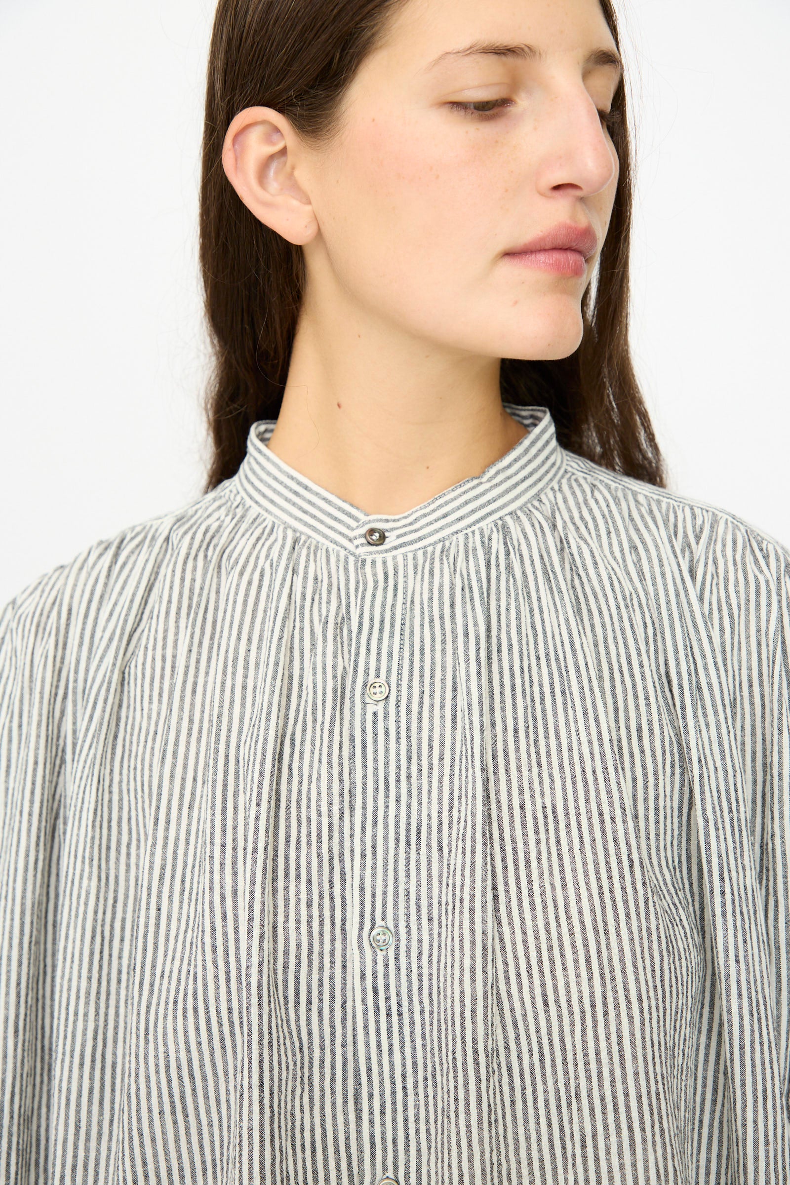 A person with long brown hair wears the Ichi Antiquités Linen and Cotton Dress in White Stripe, featuring a high collar, button-up front, and vertical grey and white stripes, looking slightly to the side.