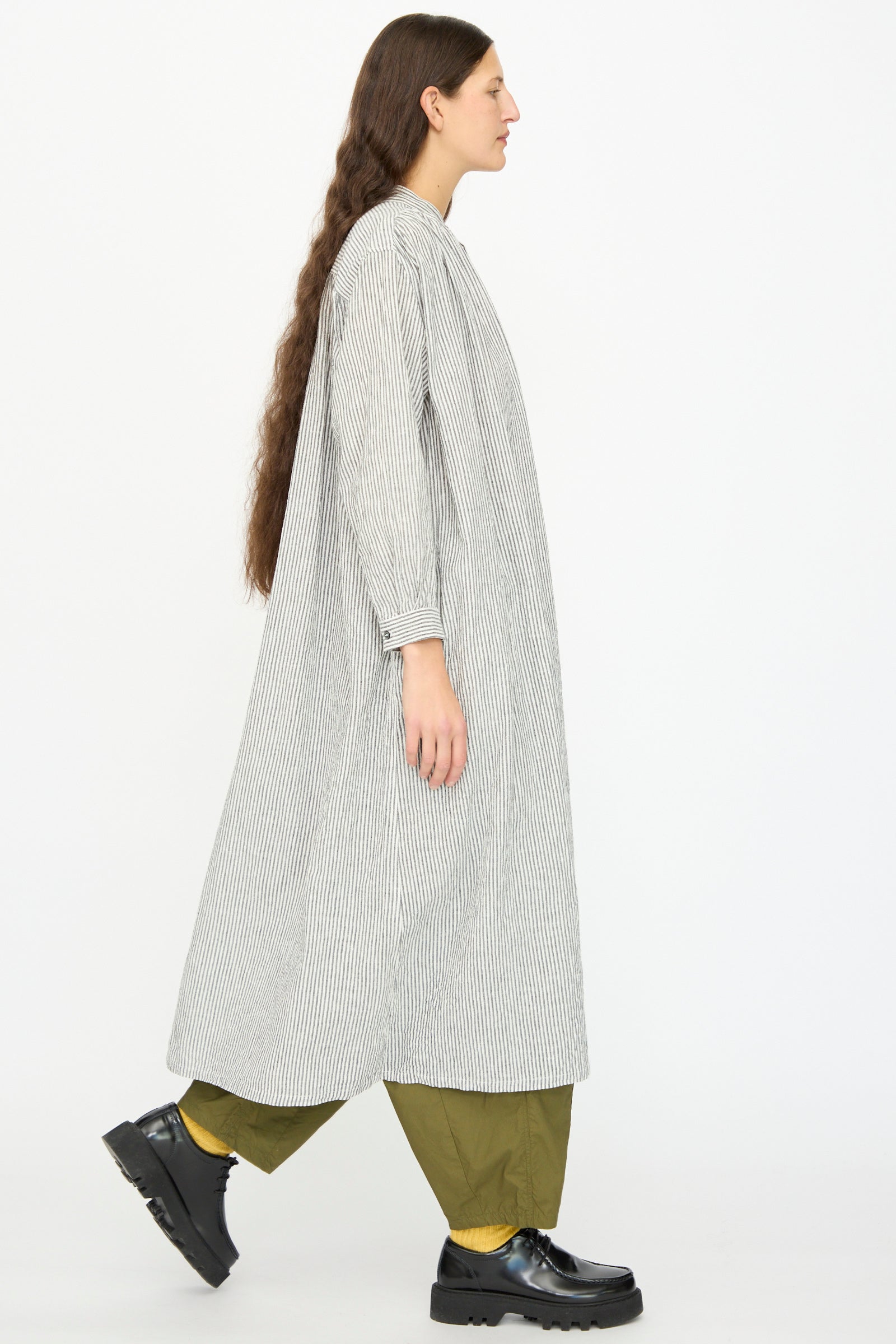 A person with long hair walks in profile wearing the Ichi Antiquités Linen and Cotton Dress in White Stripe over olive green pants, yellow socks, and black shoes against a plain white background.