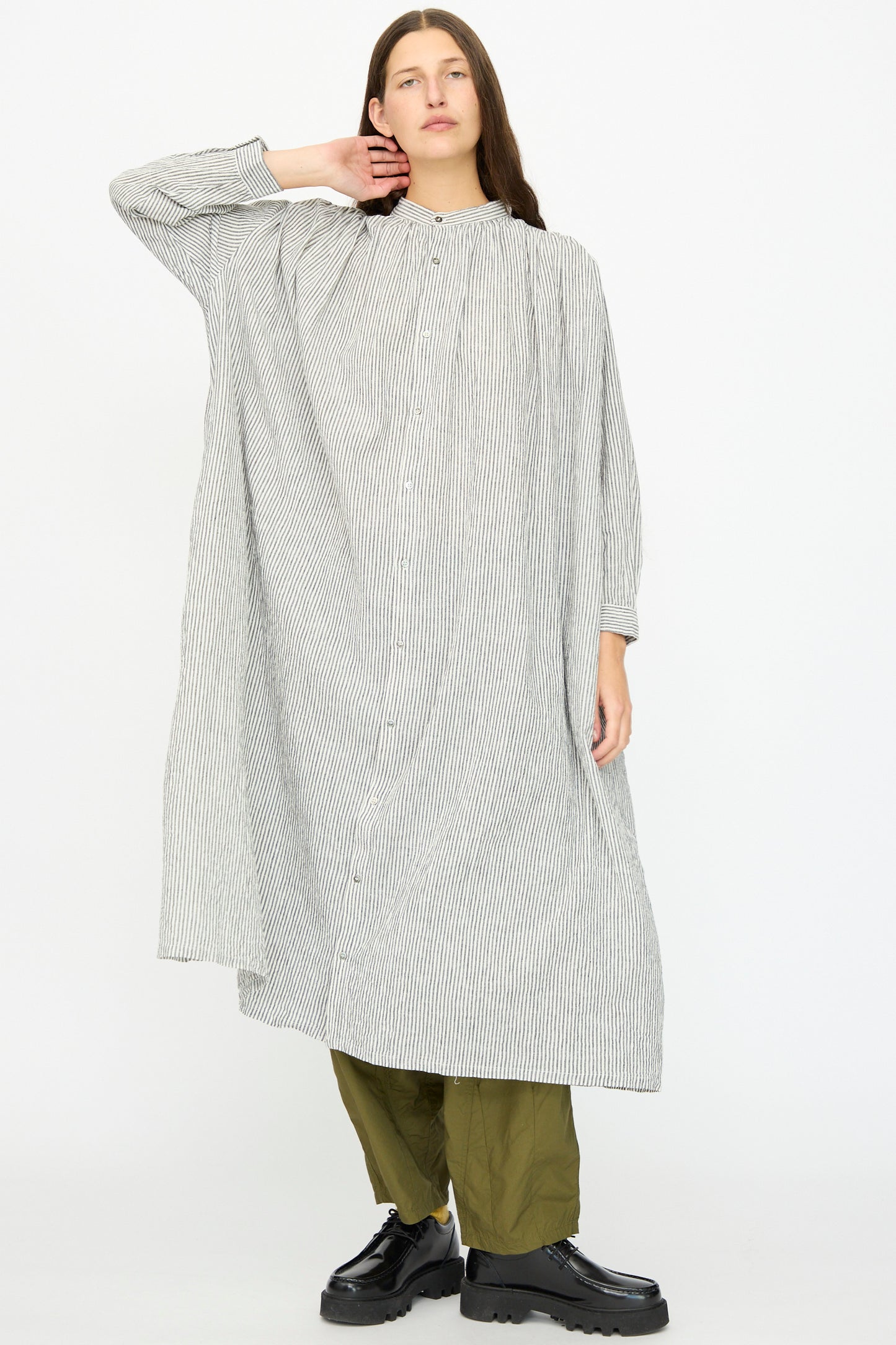 A person wears the Ichi Antiquités Linen and Cotton Dress in White Stripe over olive green pants and black platform shoes, standing against a plain light background.