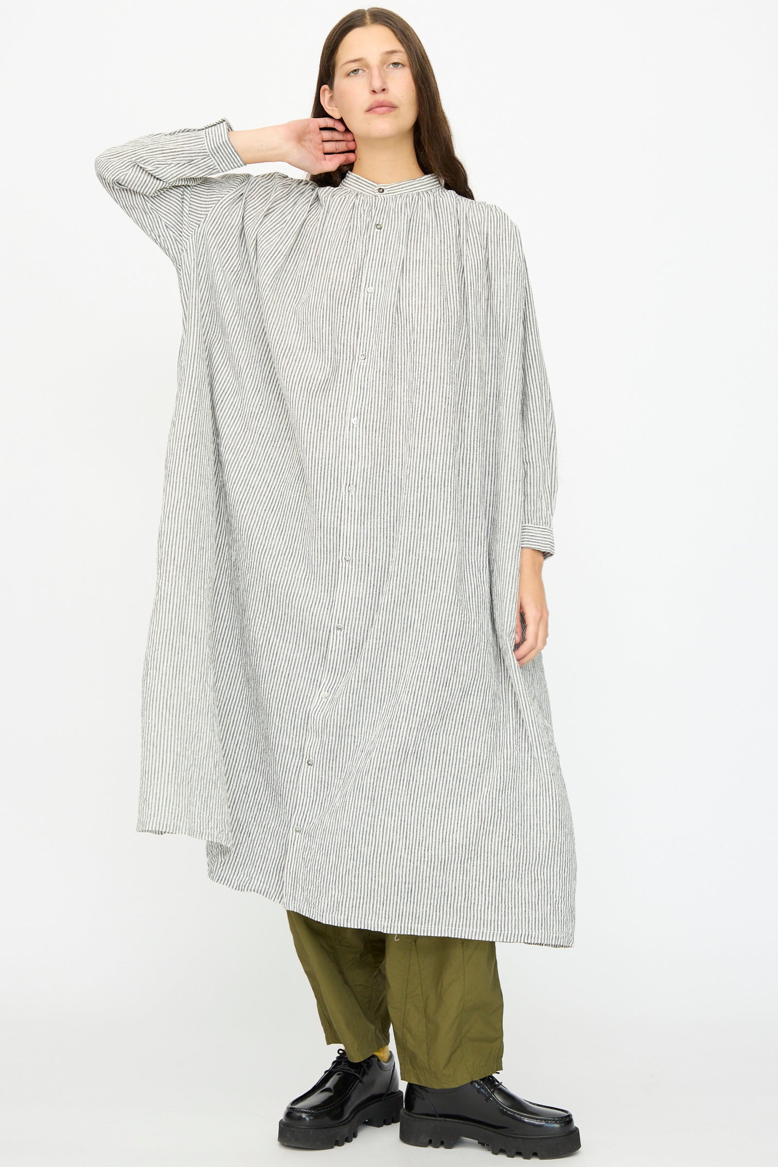 A person wears the Ichi Antiquités Linen and Cotton Dress in White Stripe over olive green pants and black platform shoes, standing against a plain light background.