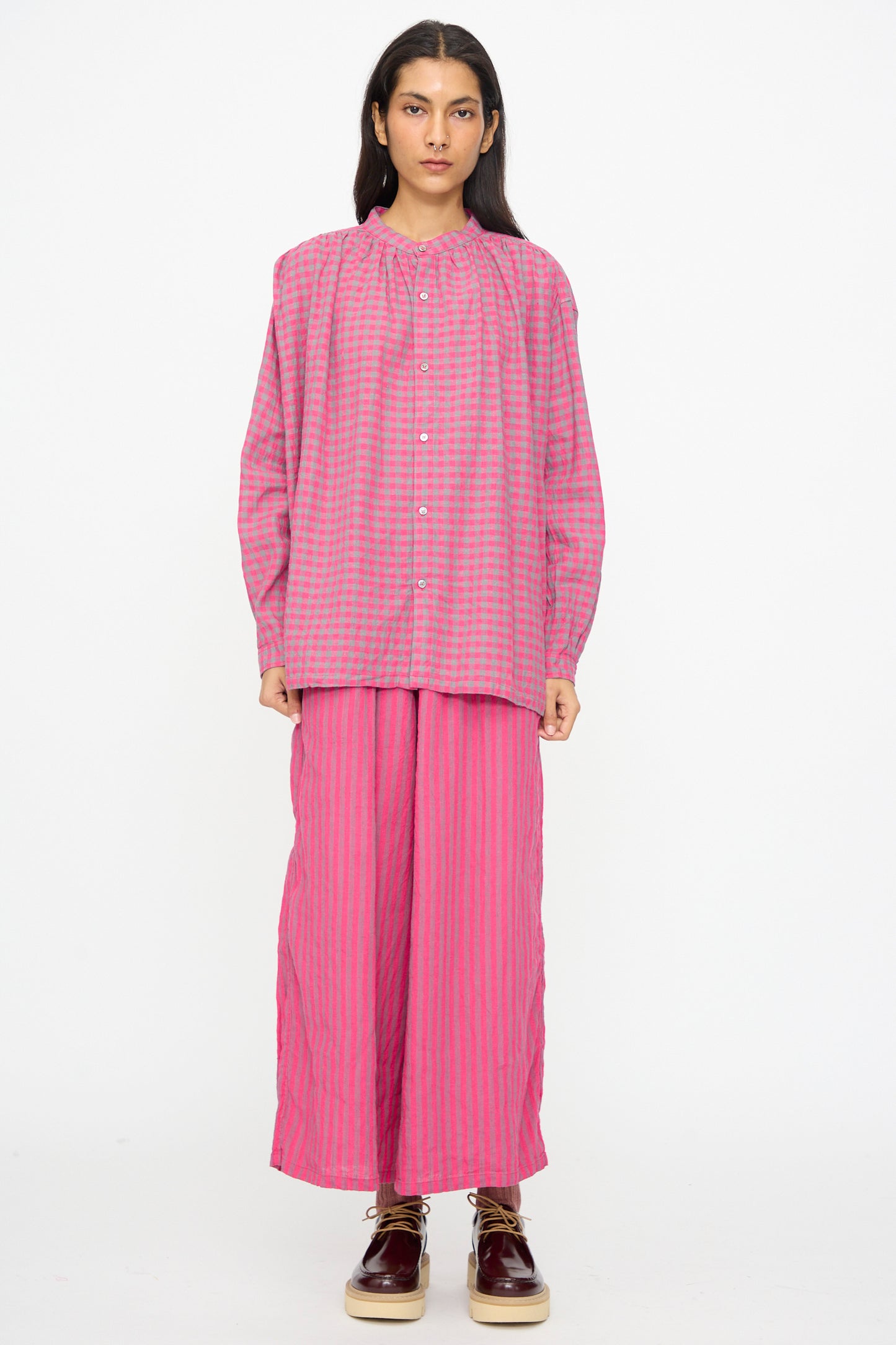 A person stands facing forward against a plain white background, wearing the Ichi Antiquités Linen Cotton Gingham Check Shirt in Pink, styled loosely with matching wide-leg pants and brown shoes.