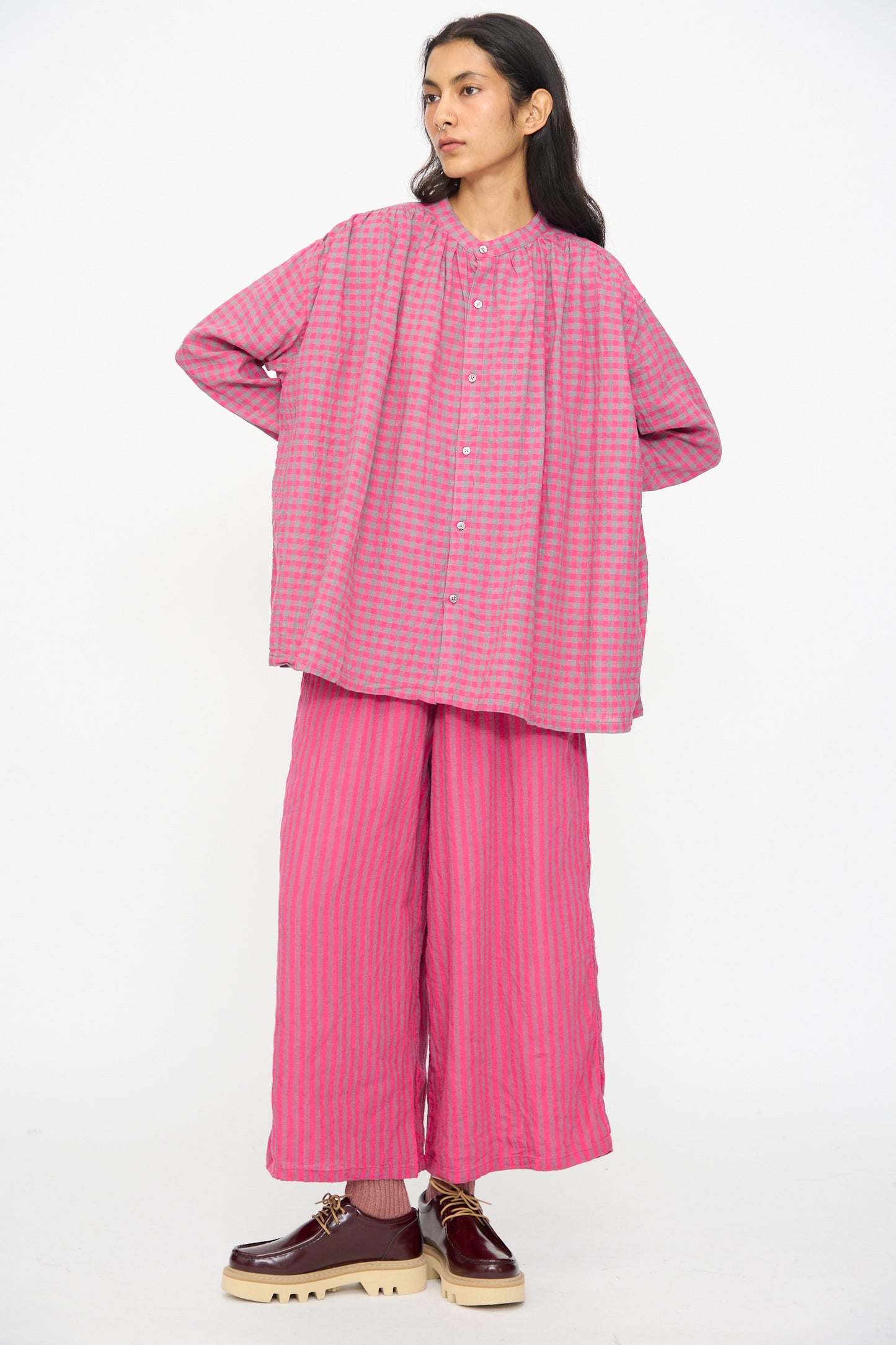 A person stands against a white background wearing the Ichi Antiquités Linen Cotton Gingham Check Shirt in Pink, styled with wide-leg pink striped pants and brown platform shoes.