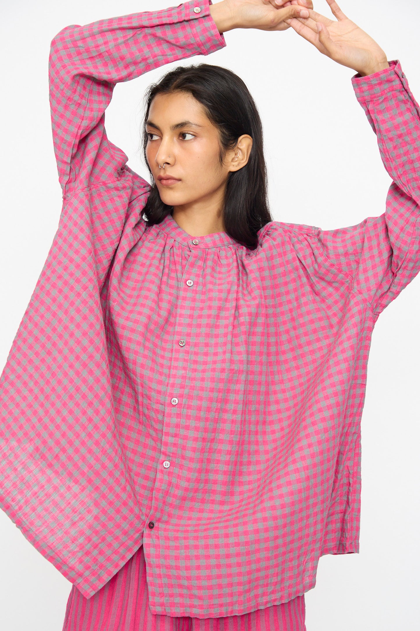 A person wearing the Ichi Antiquités Linen Cotton Gingham Check Shirt in Pink, featuring long sleeves, a band collar, and a breathable linen-cotton blend, stands with arms raised against a plain background.