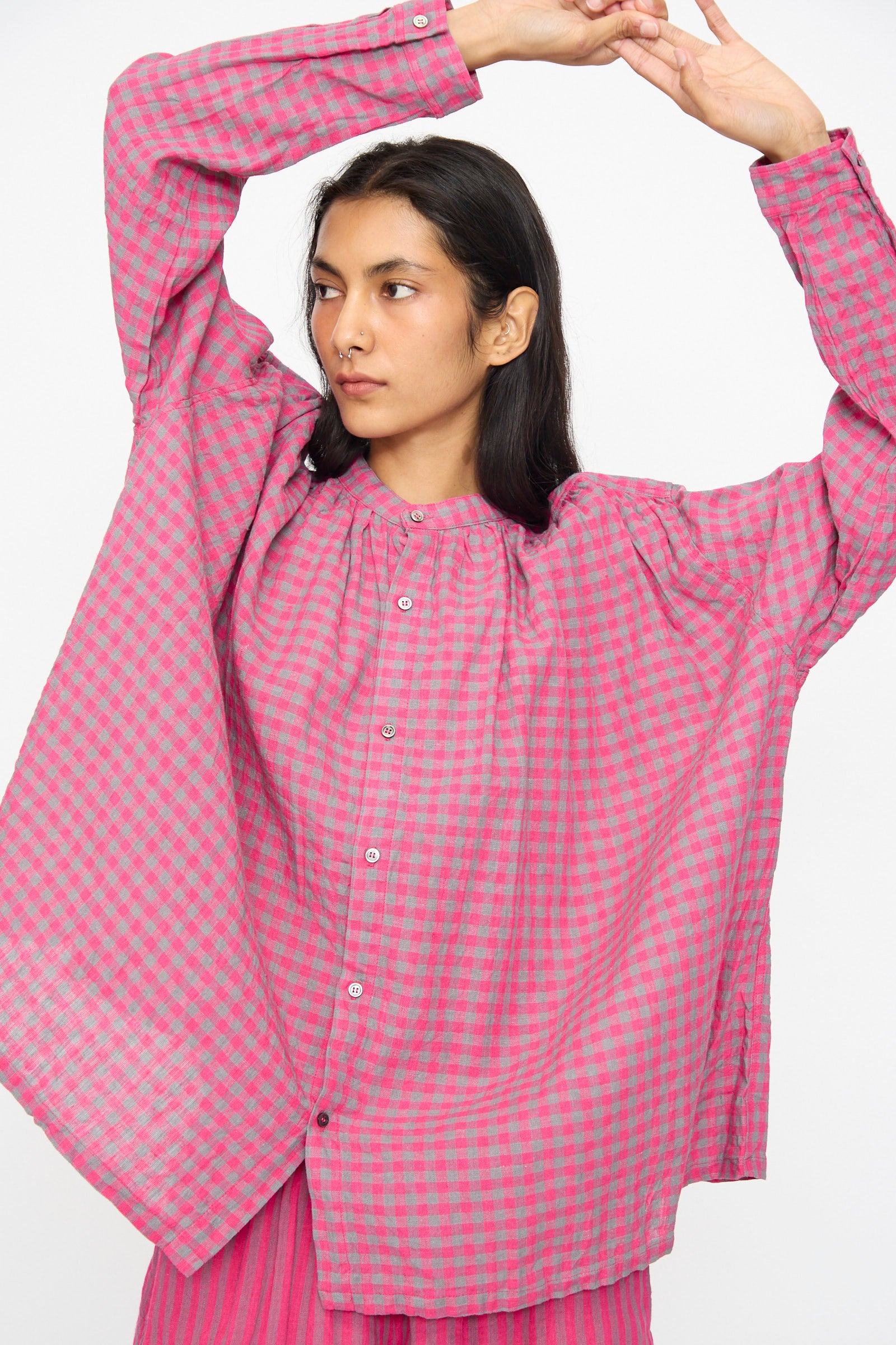 A person wearing the Ichi Antiquités Linen Cotton Gingham Check Shirt in Pink, featuring long sleeves, a band collar, and a breathable linen-cotton blend, stands with arms raised against a plain background.