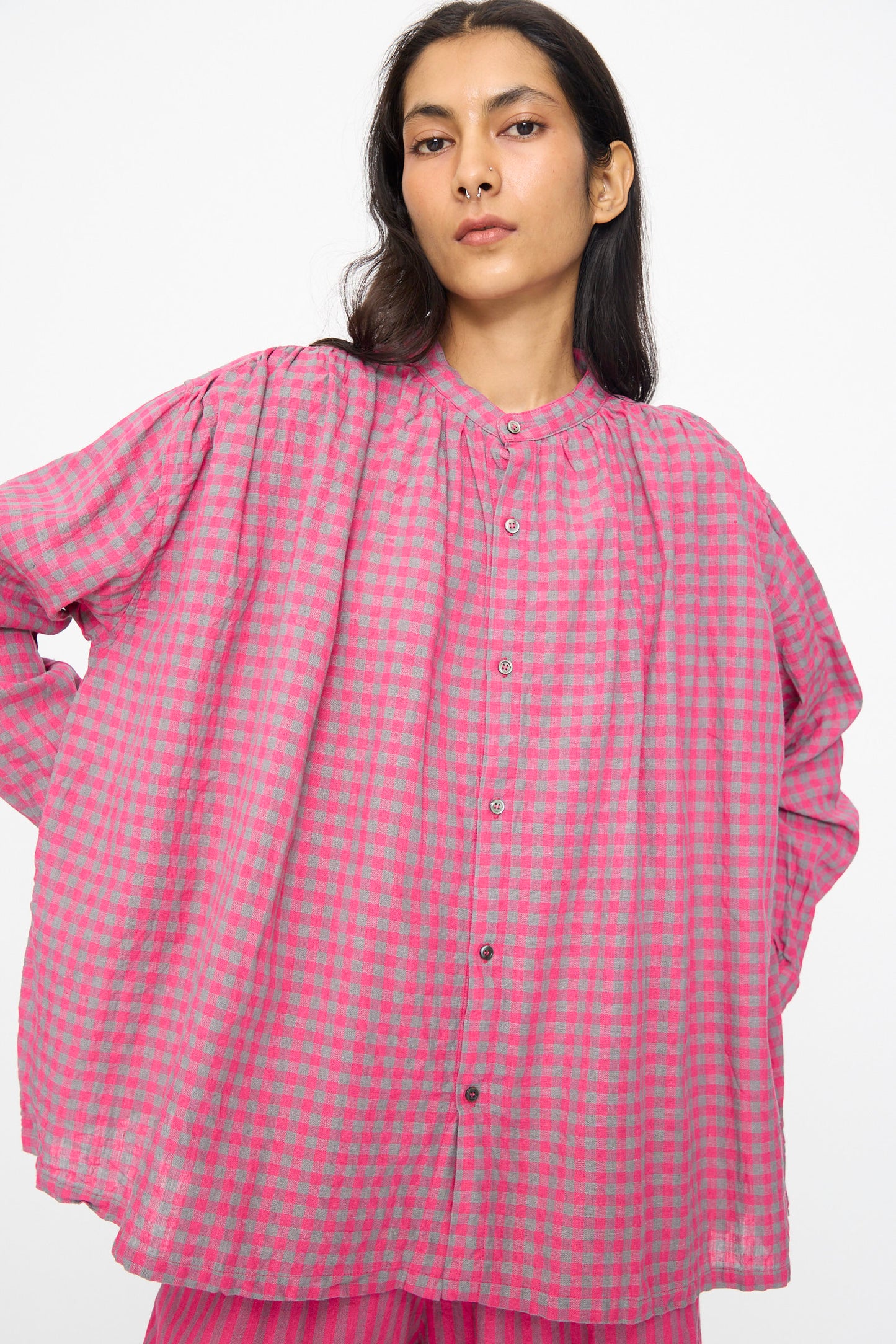 A person wears the Ichi Antiquités Linen Cotton Gingham Check Shirt in Pink, featuring an oversized fit and band collar, standing hands-on-hips against a plain white background.