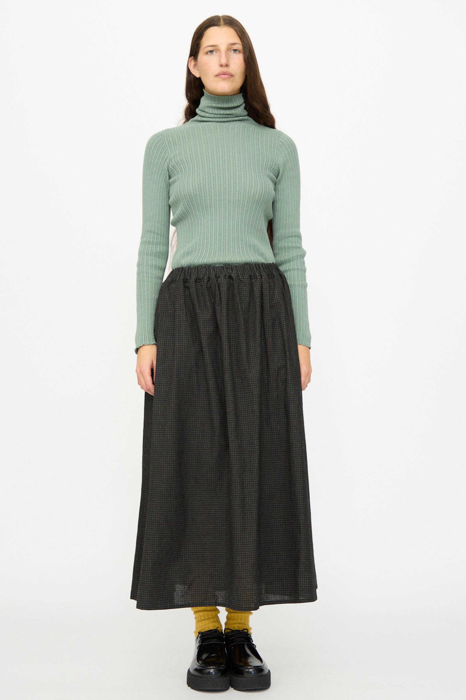 A woman stands facing forward against a white background, wearing a ribbed green turtleneck, yellow socks, black shoes, and the Ichi Antiquités Linen Cotton Gingham Sumi Skirt in Check.