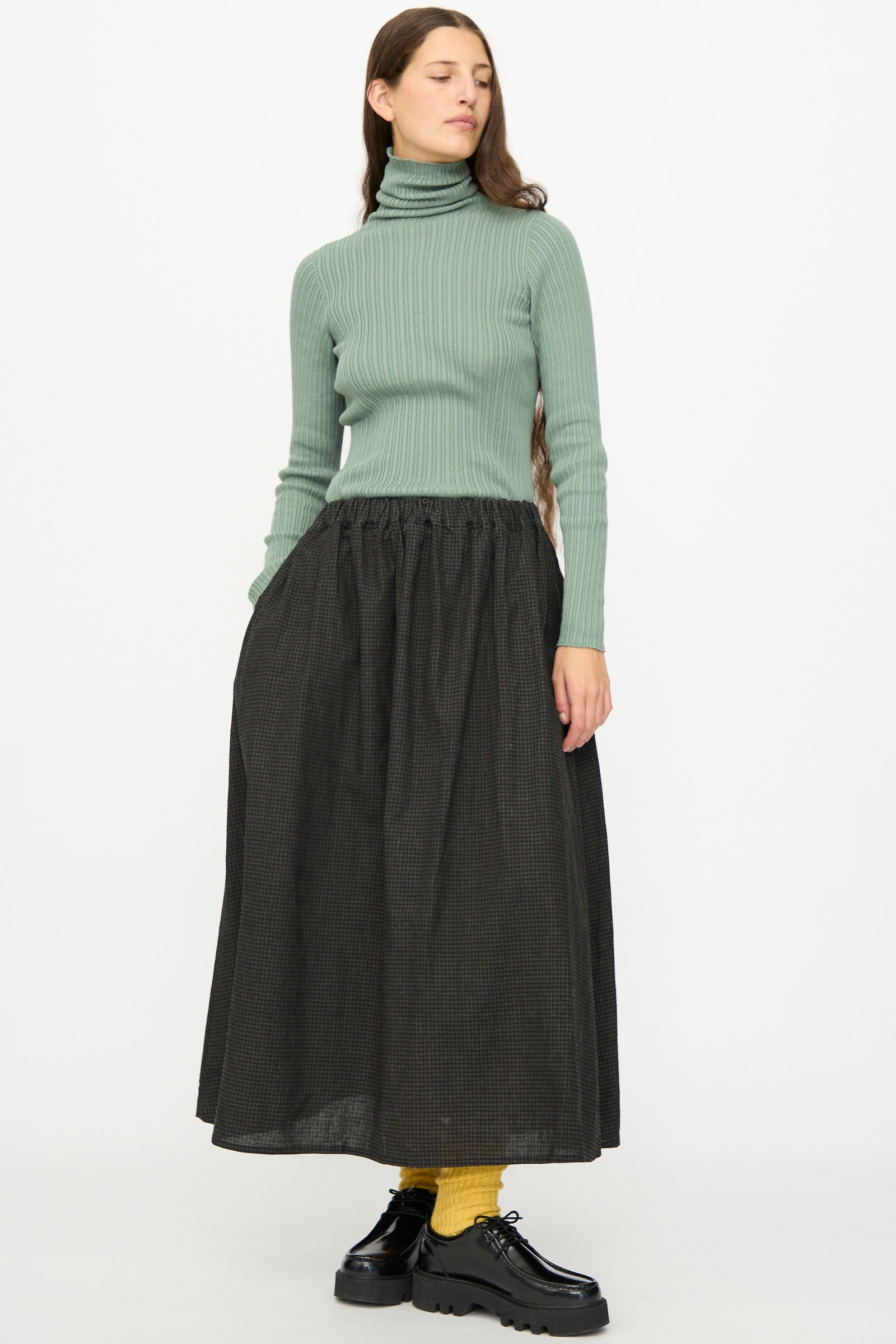 A woman stands against a plain white background wearing a green ribbed turtleneck, yellow socks, black shoes, and the Ichi Antiquités Linen Cotton Gingham Sumi Skirt in Check.