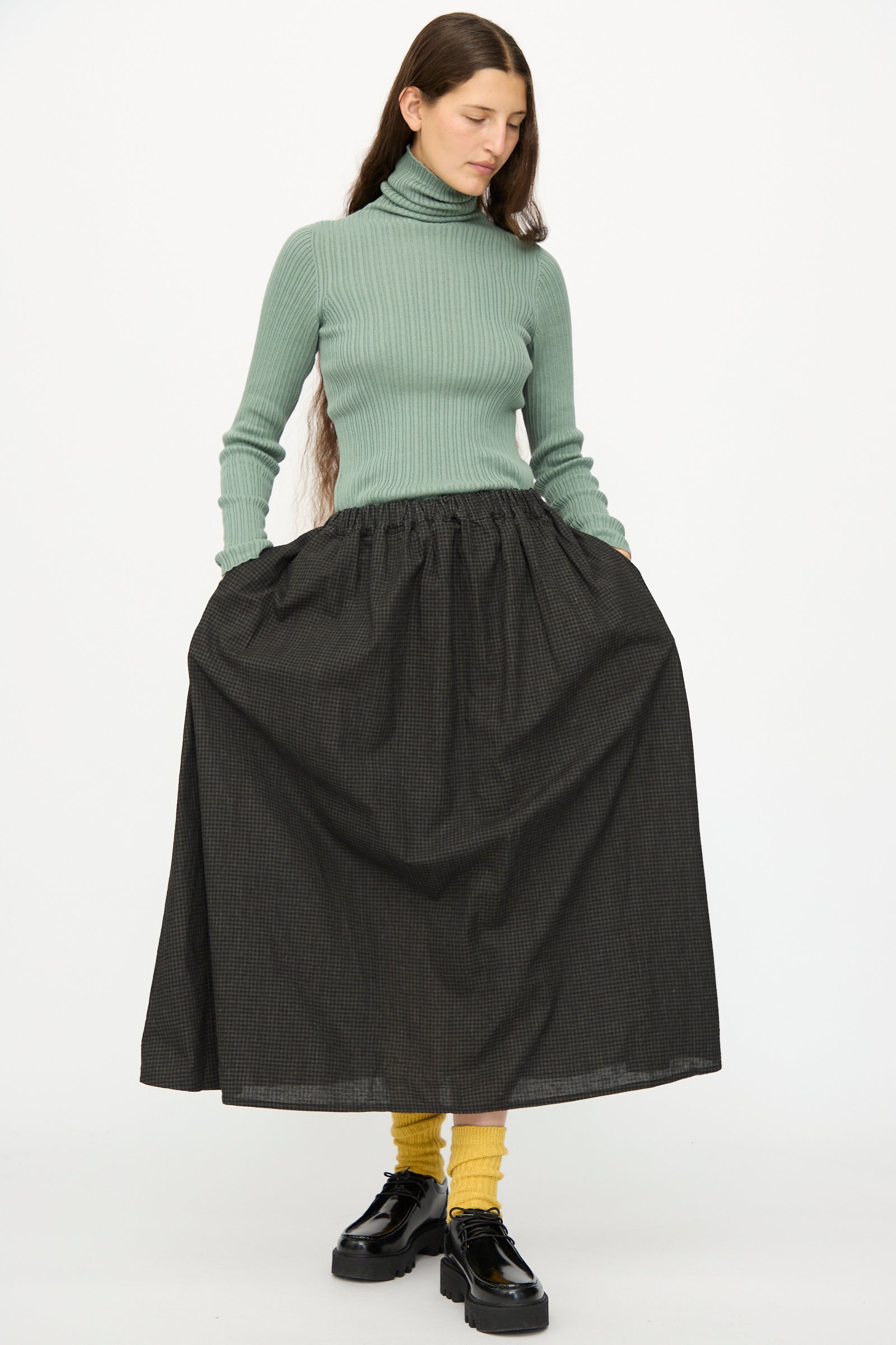 A woman wearing a light green turtleneck sweater, yellow socks, black shoes, and the Ichi Antiquités Linen Cotton Gingham Sumi Skirt in Check stands against a plain white background.