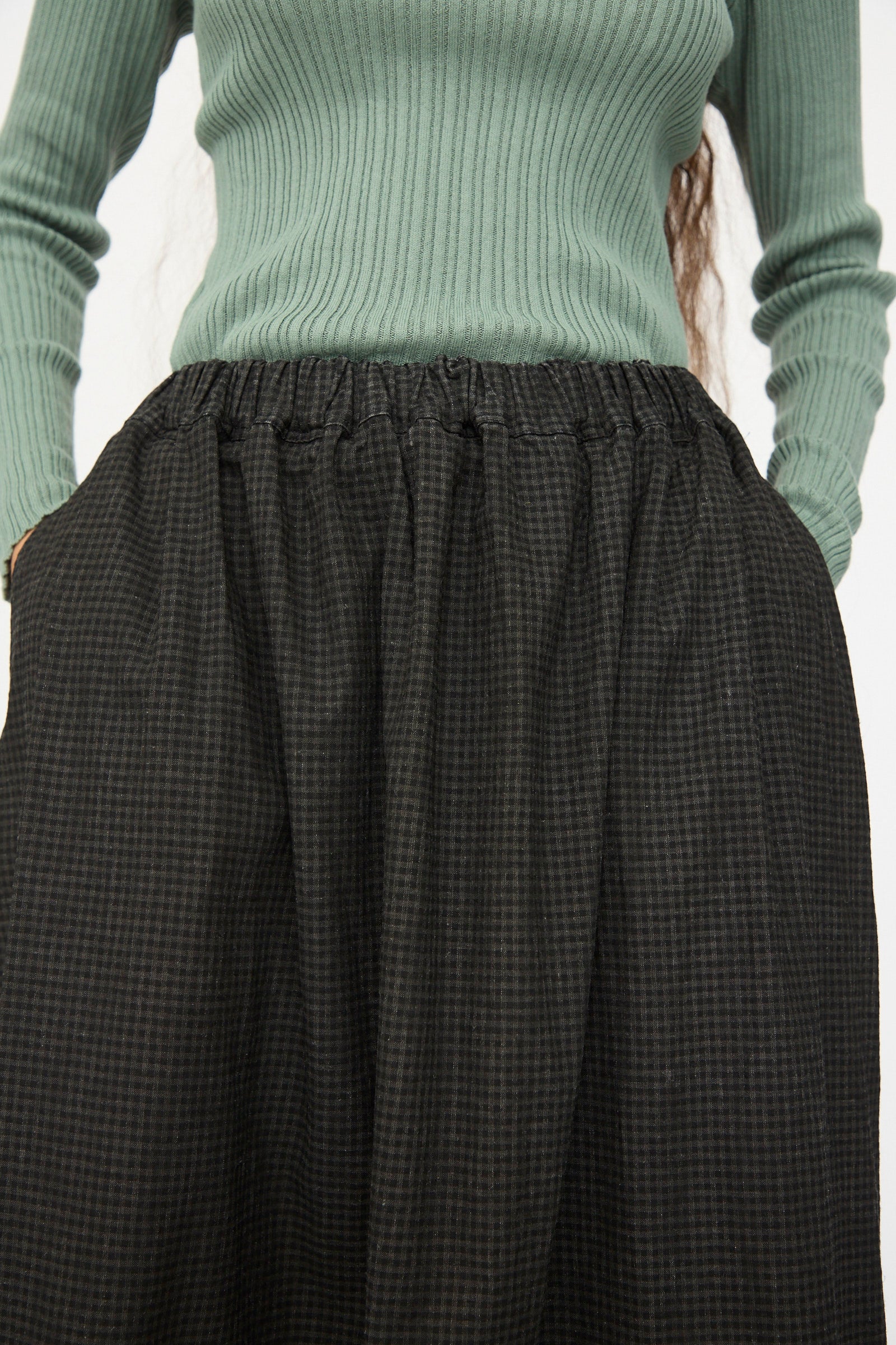 A person wearing a ribbed, long-sleeve green top and the Ichi Antiquités Linen Cotton Gingham Sumi Skirt in Check, styled high-waisted and A-line with hands in the skirt pockets.