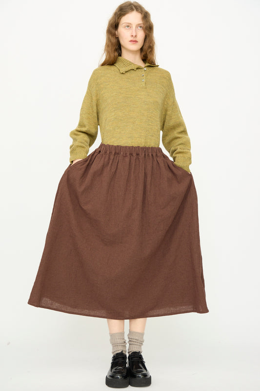 A person stands against a plain background wearing a green sweater, beige socks, black shoes, and the Ichi Antiquités Linen Cotton Houndstooth Skirt in Brown, hands tucked in skirt pockets.