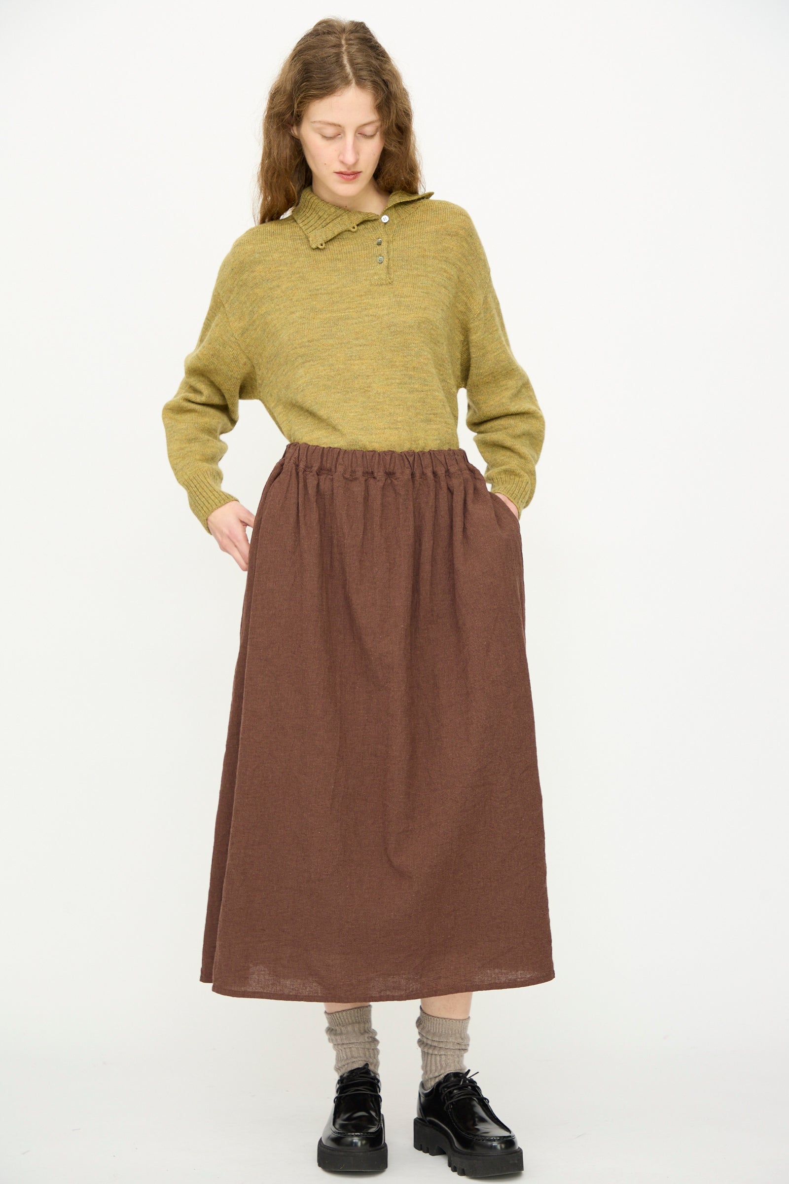 A person stands against a plain background wearing a greenish-yellow sweater, gray socks, black shoes, and the Linen Cotton Houndstooth Skirt in Brown by Ichi Antiquités, with hands tucked into the skirt’s pockets.