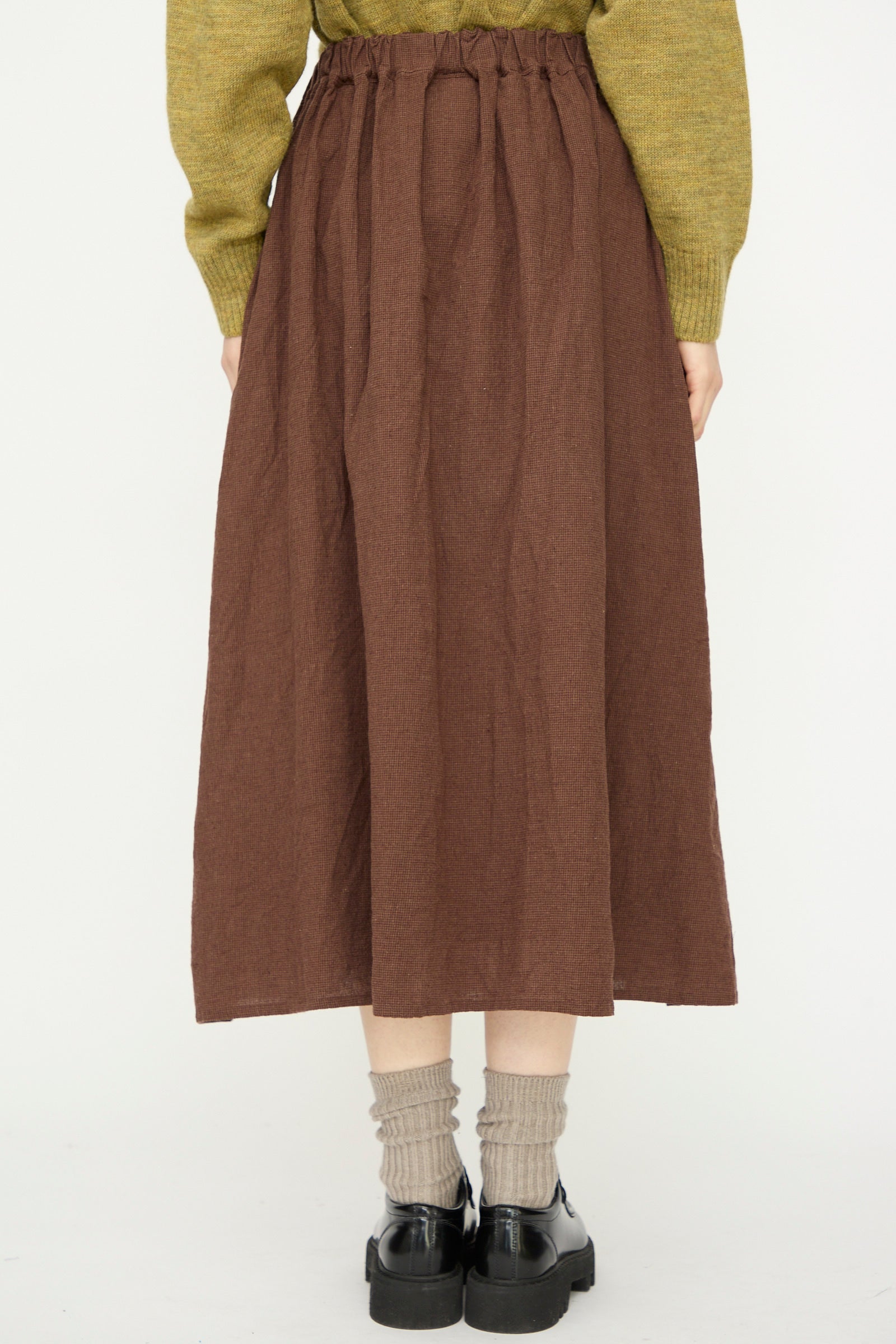 Person wearing the Ichi Antiquités Linen Cotton Houndstooth Skirt in Brown with a green sweater, beige socks, and black shoes, standing against a plain white background.