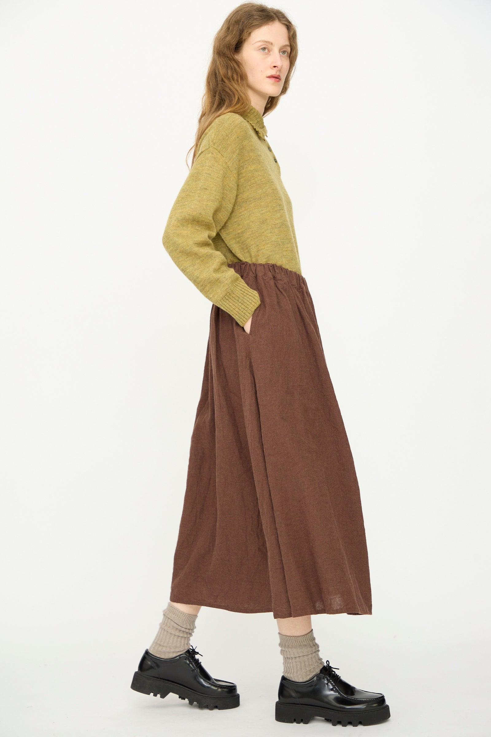 A person stands sideways against a white background wearing a green sweater, beige socks, black shoes, and the Linen Cotton Houndstooth Skirt in Brown by Ichi Antiquités.