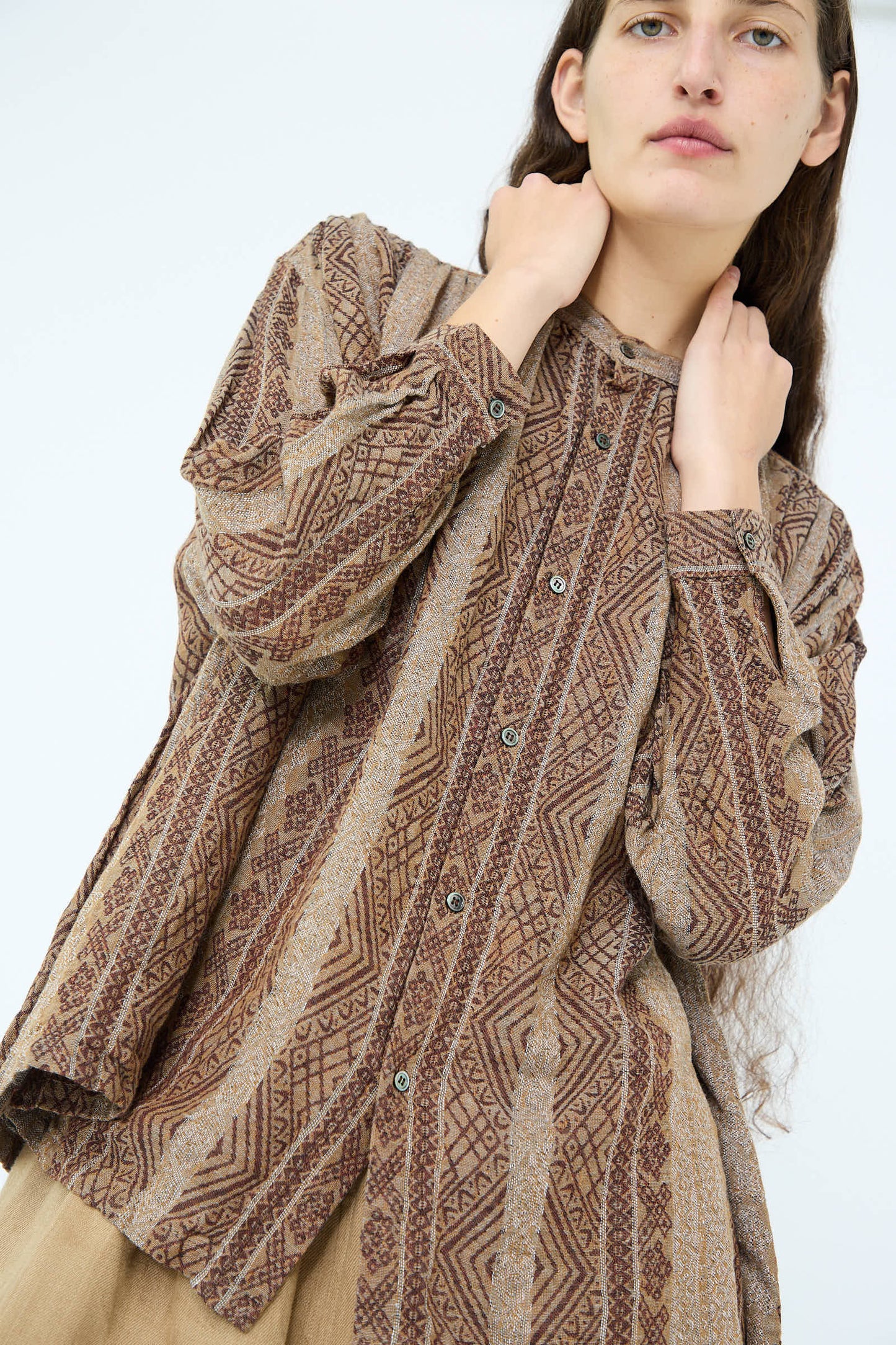 An individual is softly adjusting the band collar of a Linen Cotton Jacquard Shirt in Brown, adorned with a geometric design and long sleeves, from Ichi Antiquités.