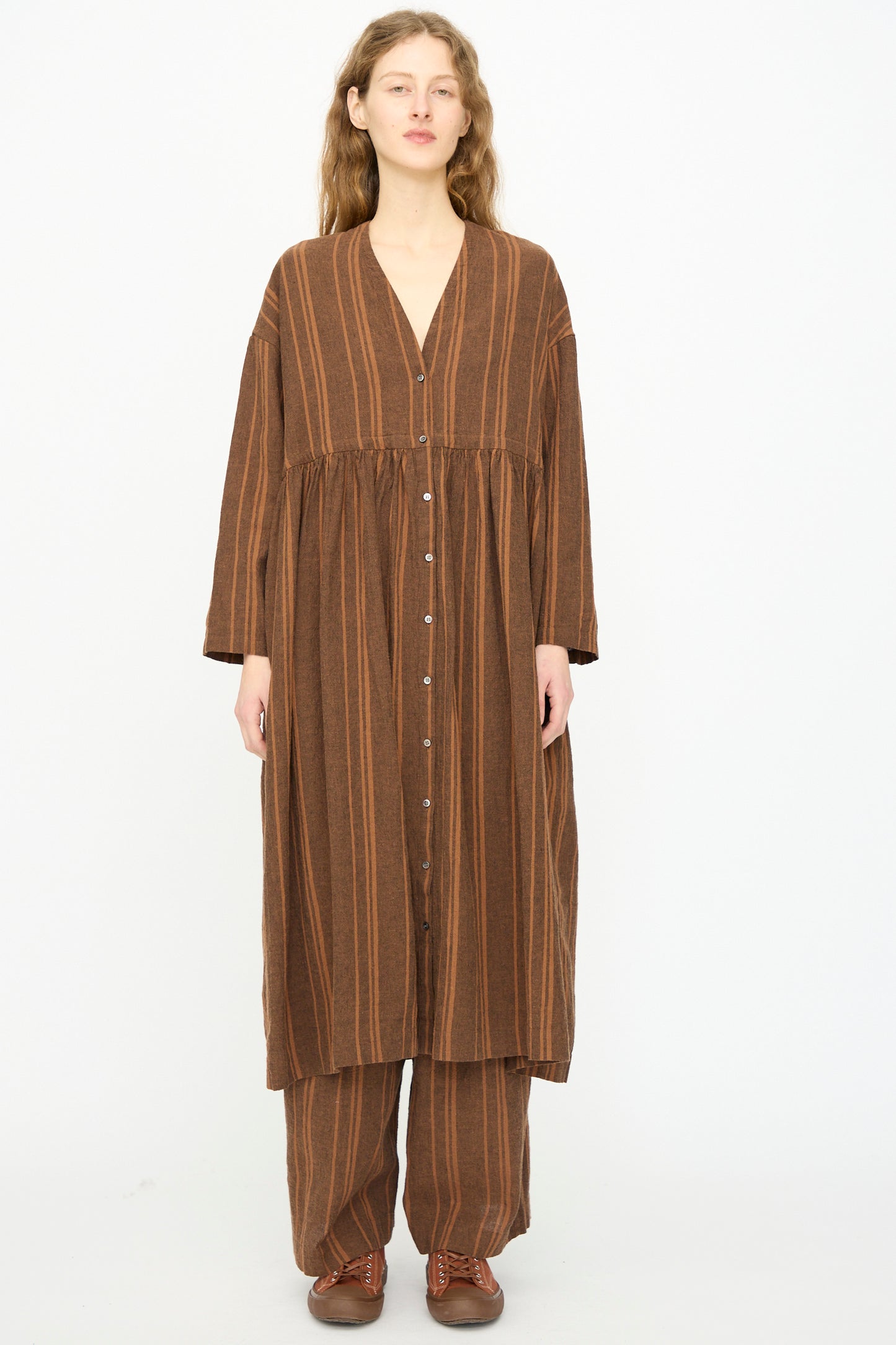 A person faces forward against a white background, wearing the Ichi Antiquités Linen Cotton Random Stripe Dress in Brown over matching wide-leg pants and brown shoes.