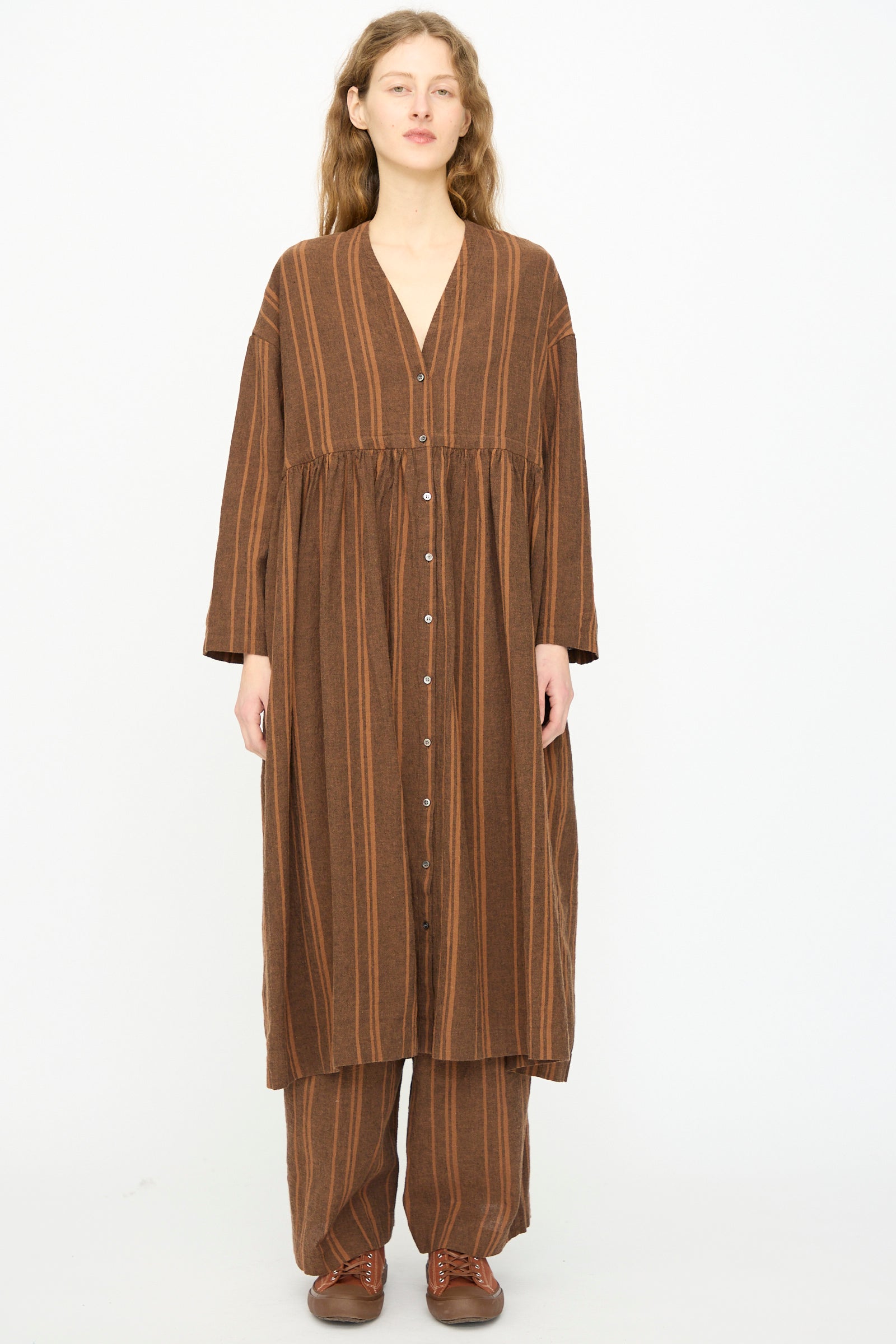 A person faces forward against a white background, wearing the Ichi Antiquités Linen Cotton Random Stripe Dress in Brown over matching wide-leg pants and brown shoes.