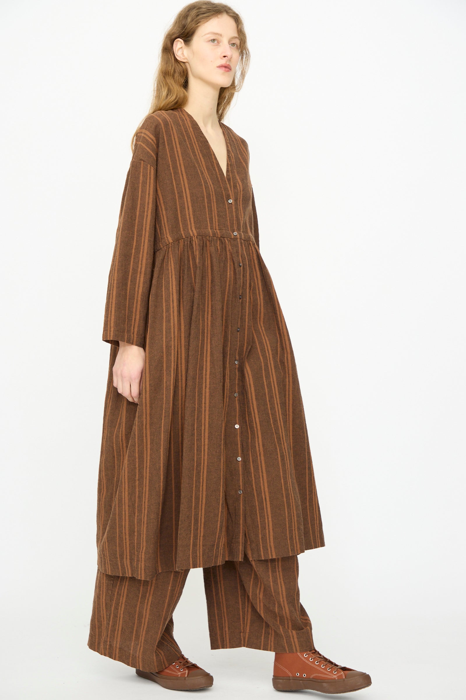 A person wearing the Ichi Antiquités Linen Cotton Random Stripe Dress in Brown stands against a plain white background, pairing it with brown lace-up shoes.