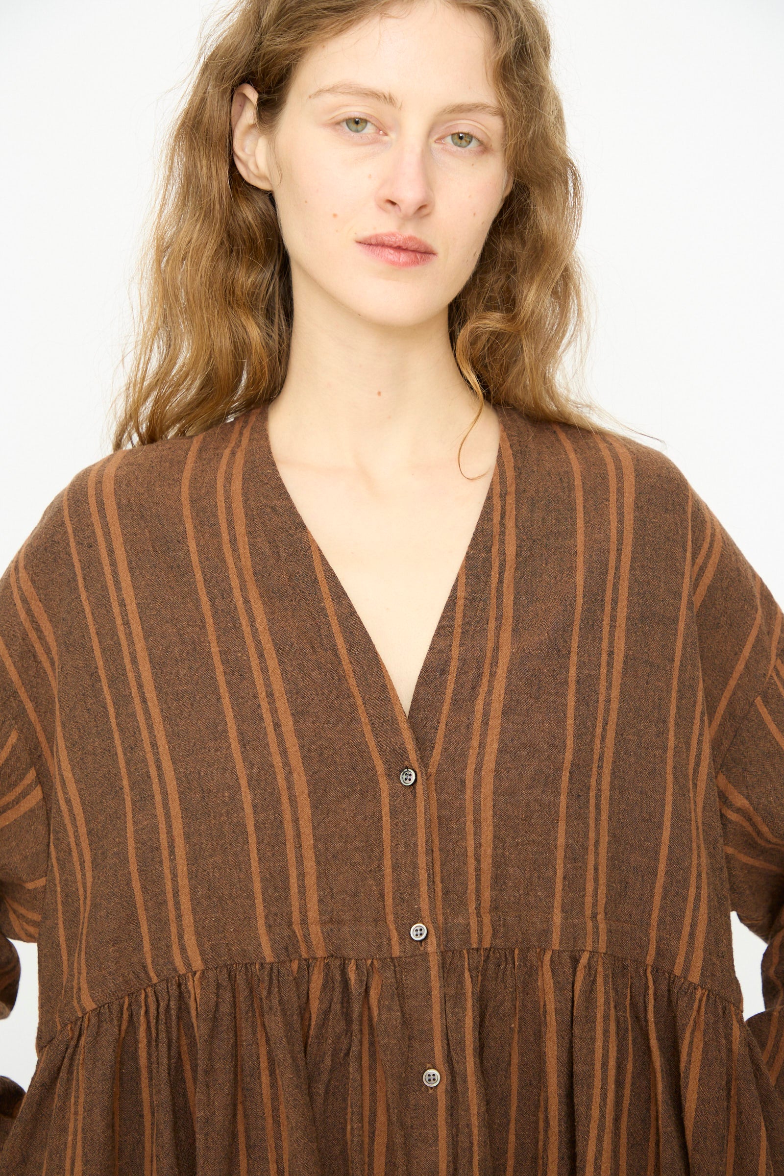 A person with long, wavy light brown hair models the Ichi Antiquités Linen Cotton Random Stripe Dress in Brown, featuring front buttons and a drawstring waist, standing against a plain white background.