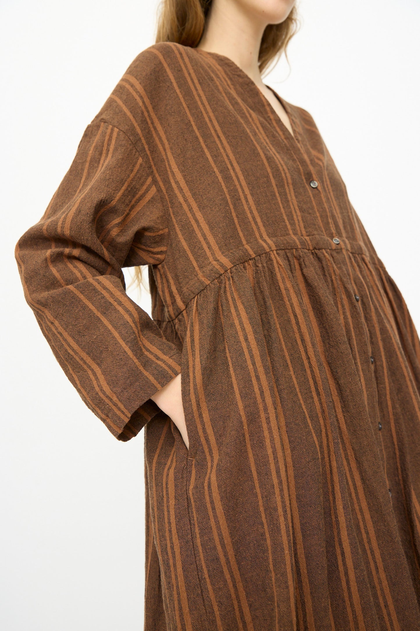 A person wears the Ichi Antiquités Linen Cotton Random Stripe Dress in Brown—long-sleeved, buttoned, with a drawstring waist and pockets—standing against a plain background.