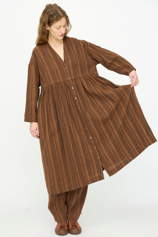A person wearing the Ichi Antiquités Linen Cotton Random Stripe Dress in Brown stands against a white background, holding out the side of the dress.