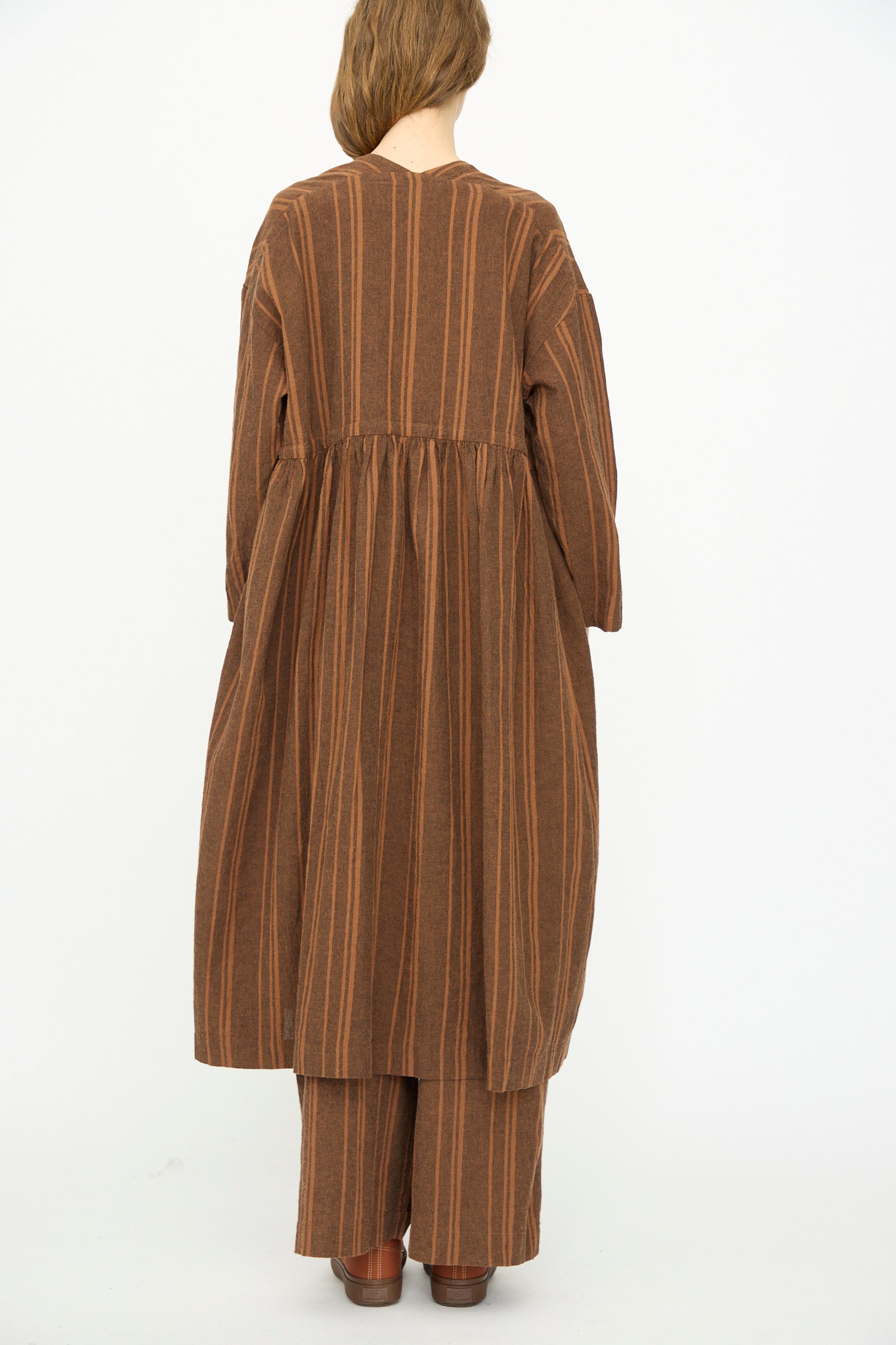A person stands with their back to the camera, wearing the Ichi Antiquités Linen Cotton Random Stripe Dress in Brown over matching wide-leg pants and brown shoes.