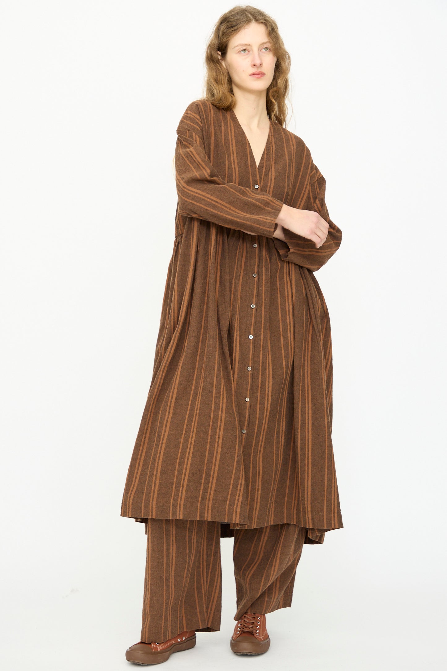 A person wears the Ichi Antiquités Linen Cotton Random Stripe Dress in Brown, styled over matching pants and brown shoes, standing against a plain white background.