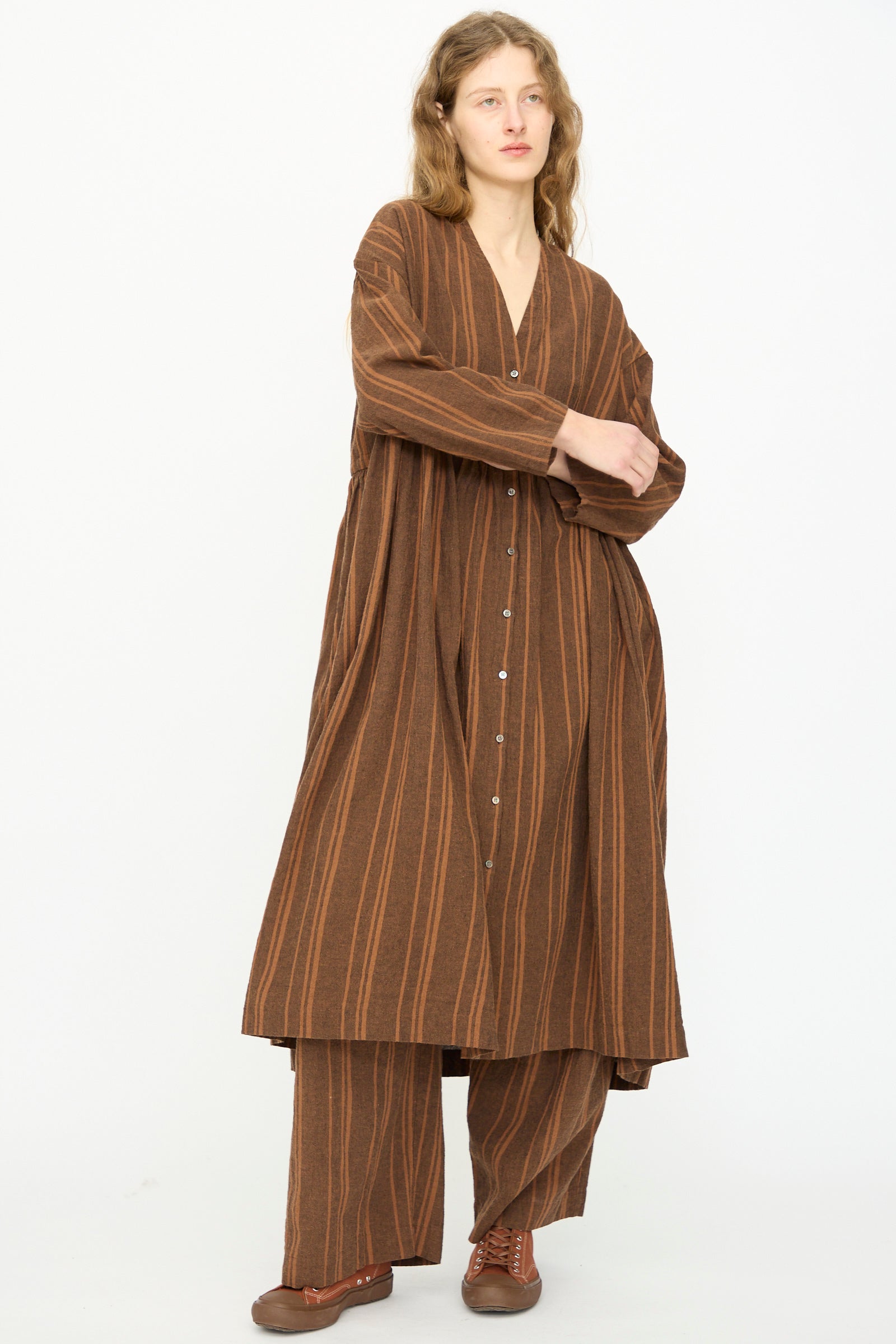 A person wears the Ichi Antiquités Linen Cotton Random Stripe Dress in Brown, styled over matching pants and brown shoes, standing against a plain white background.