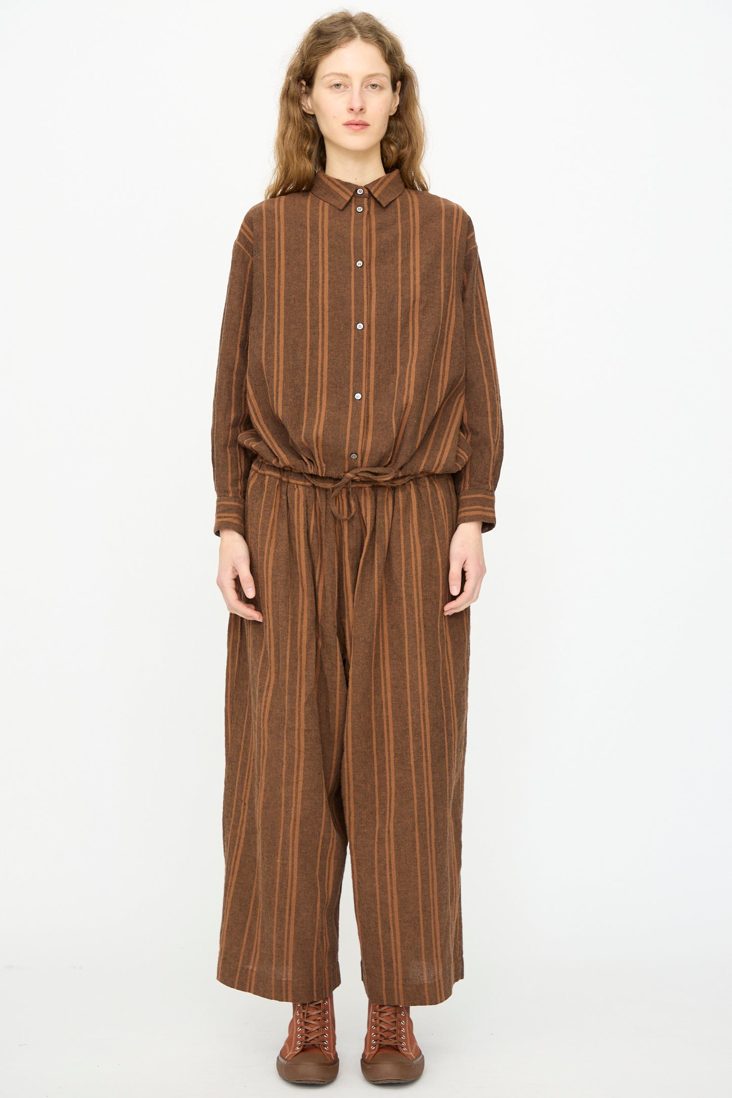 A person faces forward wearing Ichi Antiquités’ Linen Cotton Random Stripe Pant in Brown, featuring vertical stripes, elastic waist, and a relaxed fit. Paired with a matching long-sleeve shirt and brown boots. Made in Japan.