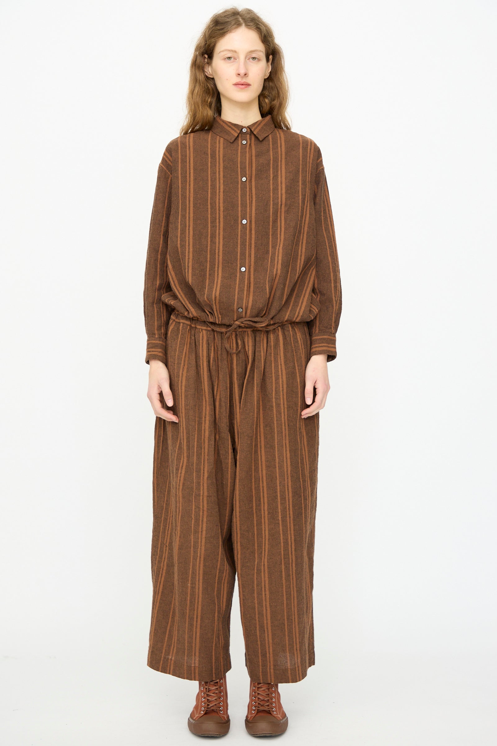 A person faces forward wearing Ichi Antiquités’ Linen Cotton Random Stripe Pant in Brown, featuring vertical stripes, elastic waist, and a relaxed fit. Paired with a matching long-sleeve shirt and brown boots. Made in Japan.