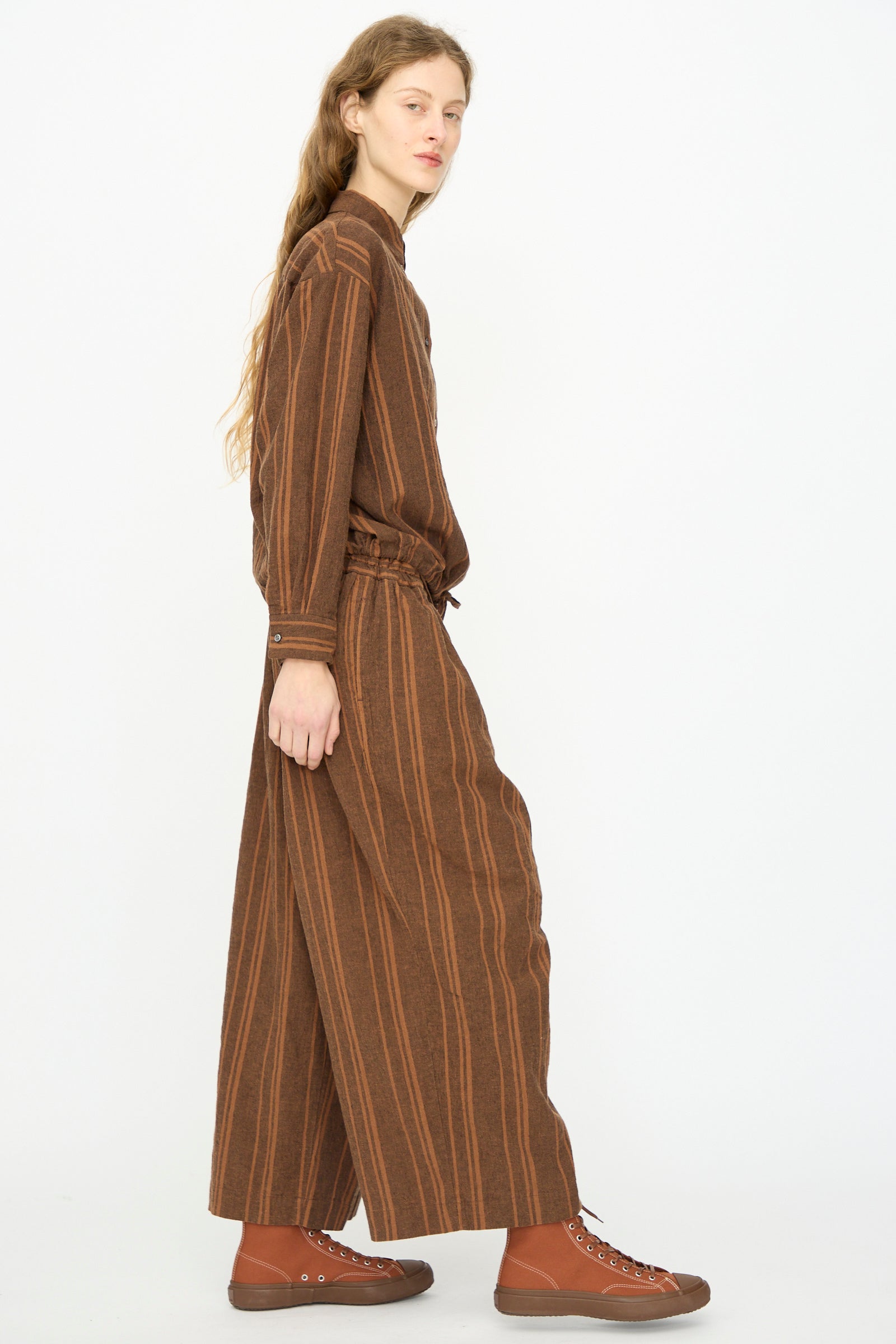 A person stands in profile against a white background, wearing Ichi Antiquités Linen Cotton Random Stripe Pant in Brown with an elastic waist and brown sneakers, paired with a matching long-sleeved striped dress.