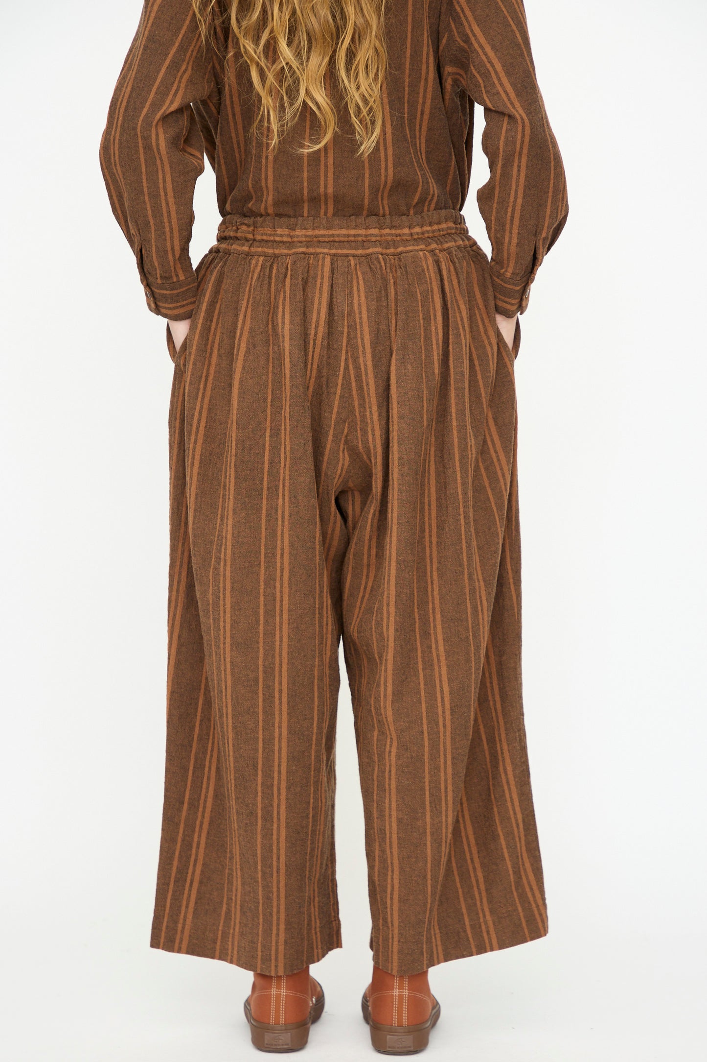 Person standing with back to camera, wearing Ichi Antiquités Linen Cotton Random Stripe Pant in Brown and brown sandals on a white background.
