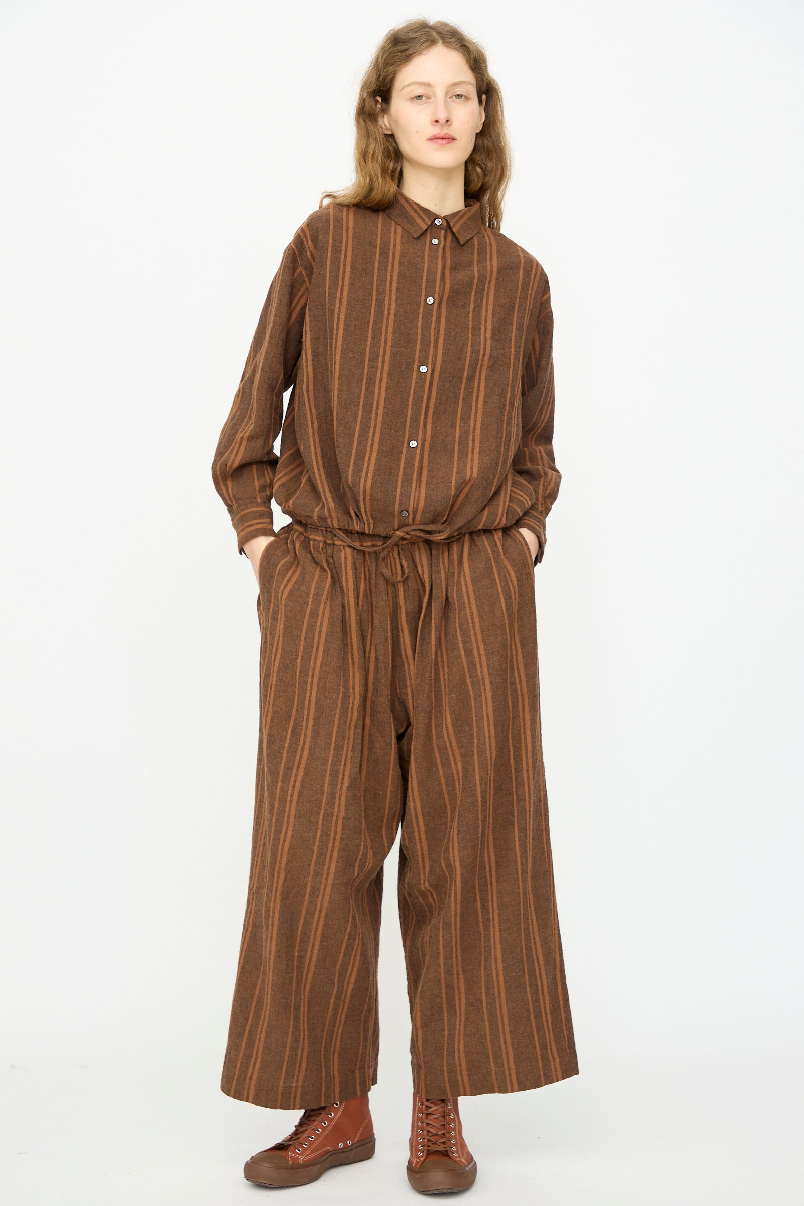 A person stands facing forward wearing Ichi Antiquités Linen Cotton Random Stripe Pant in Brown, featuring an elastic waist and brown stripes, paired with a matching striped button-up shirt and brown lace-up shoes, hands in pockets, against a plain white background.
