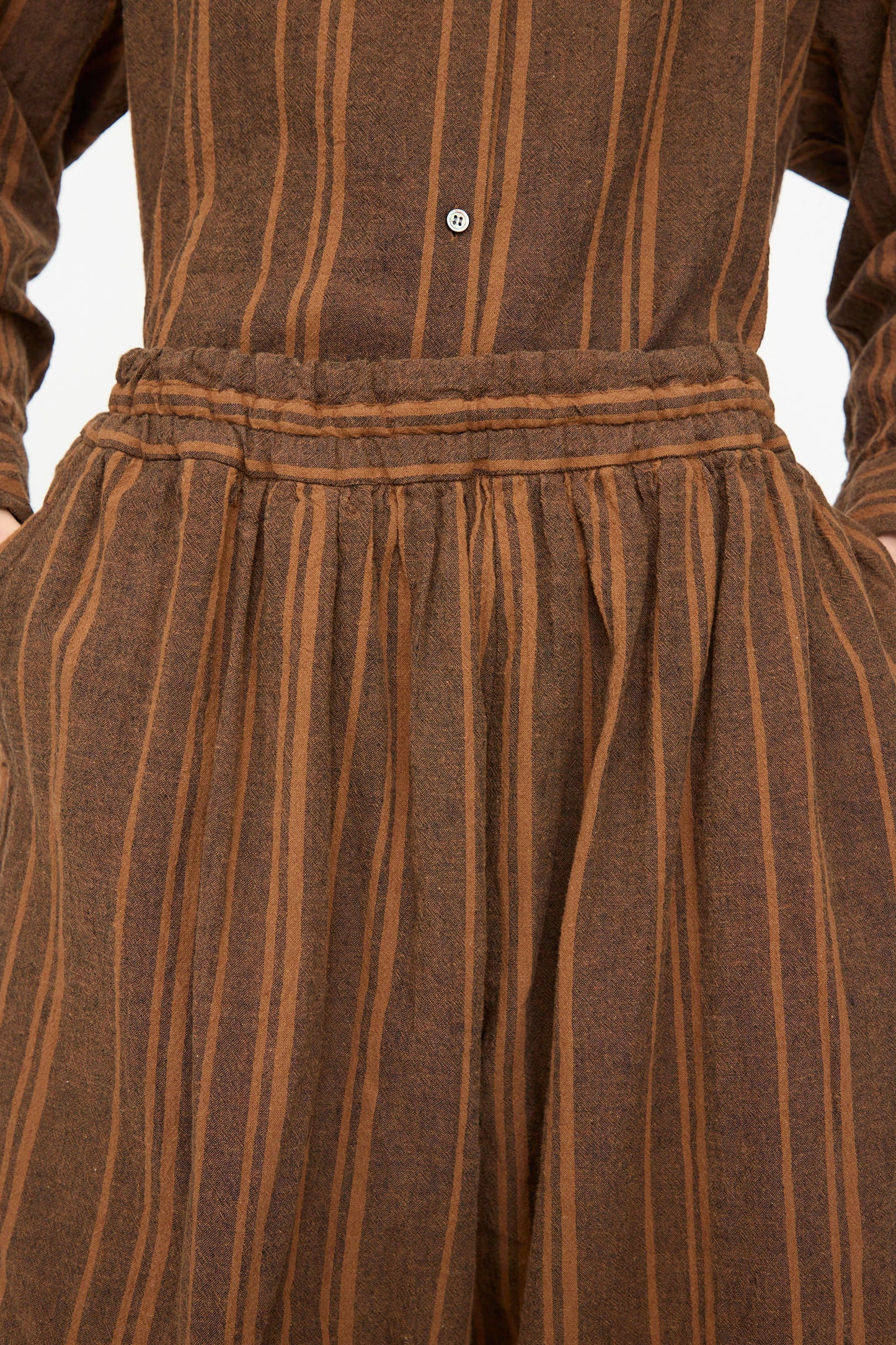 Close-up of the Ichi Antiquités Linen Cotton Random Stripe Pant in brown, featuring tan vertical stripes, an elastic gathered waistband, and hands in side pockets.