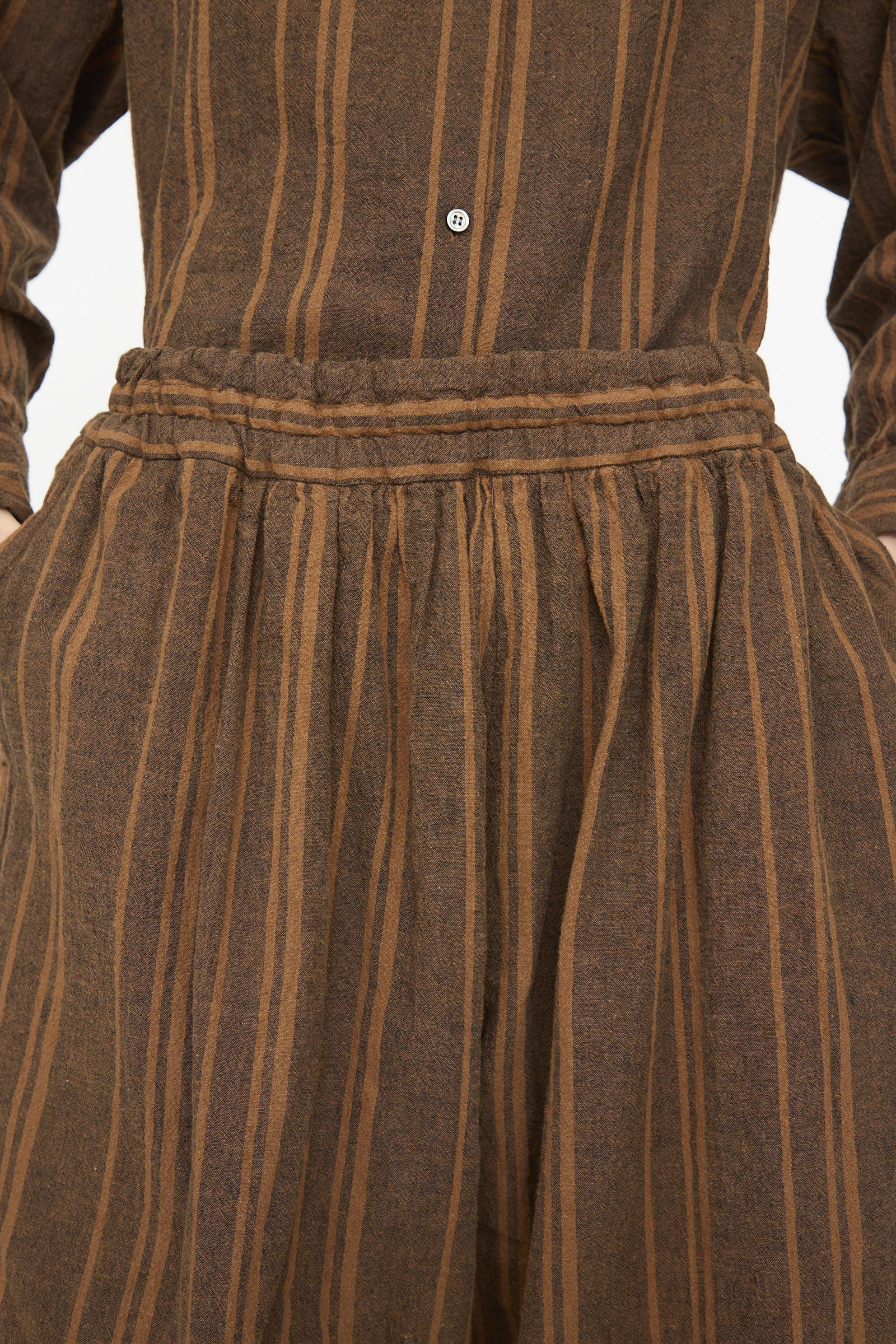 Close-up of the Ichi Antiquités Linen Cotton Random Stripe Pant in brown, featuring tan vertical stripes, an elastic gathered waistband, and hands in side pockets.