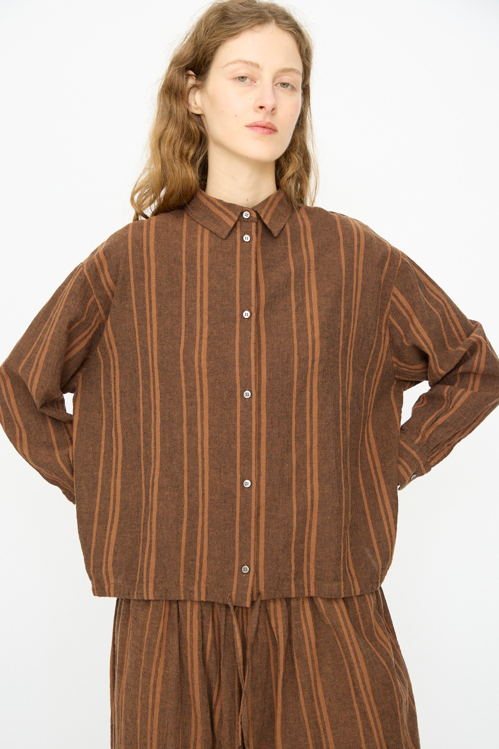 A person with long, wavy hair wears the Ichi Antiquités Linen Cotton Random Stripe Shirt in Brown, standing against a plain white background.