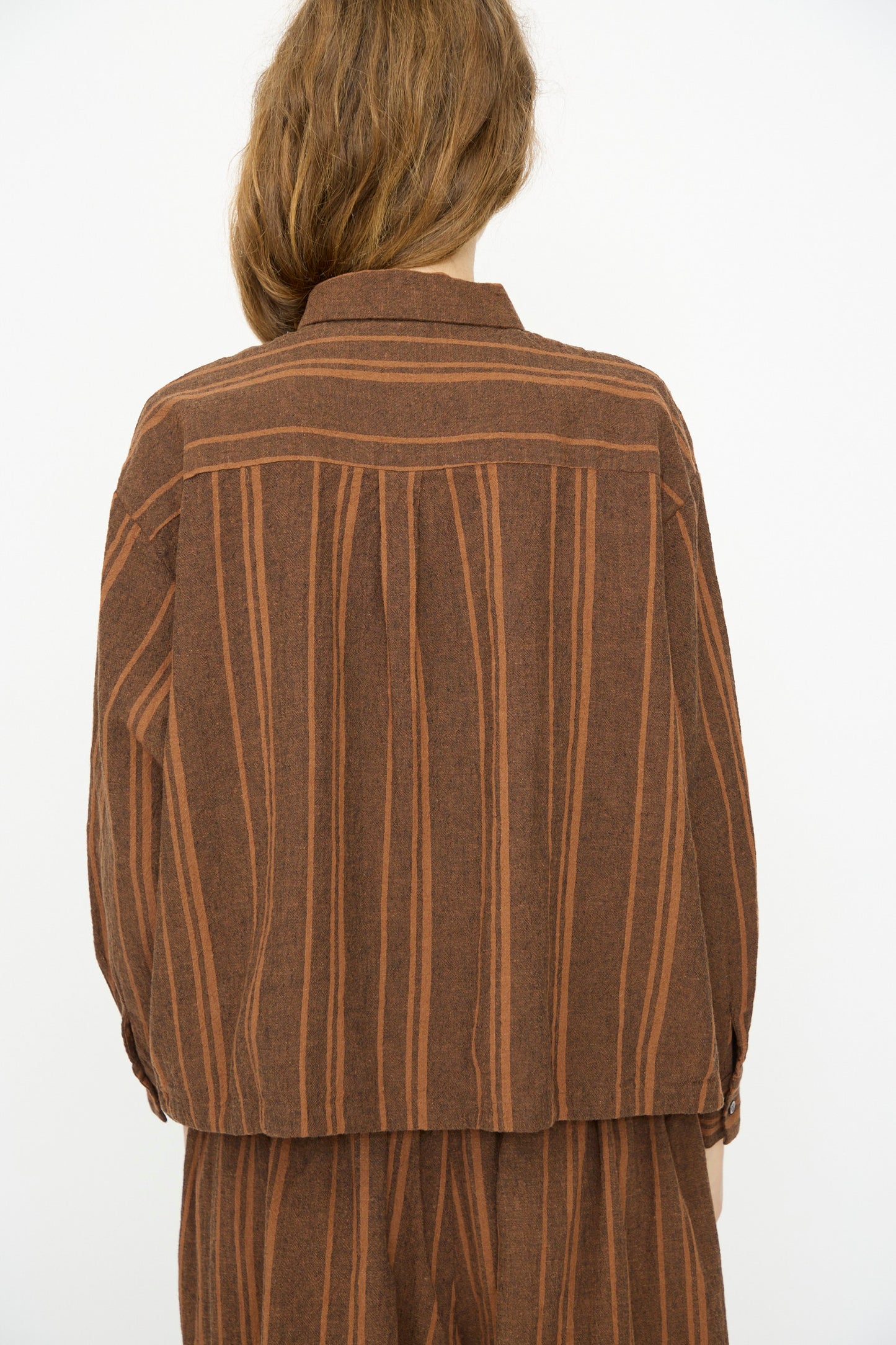 A person with long light brown hair is seen from the back wearing the Ichi Antiquités Linen Cotton Random Stripe Shirt in Brown, set against a plain white background.