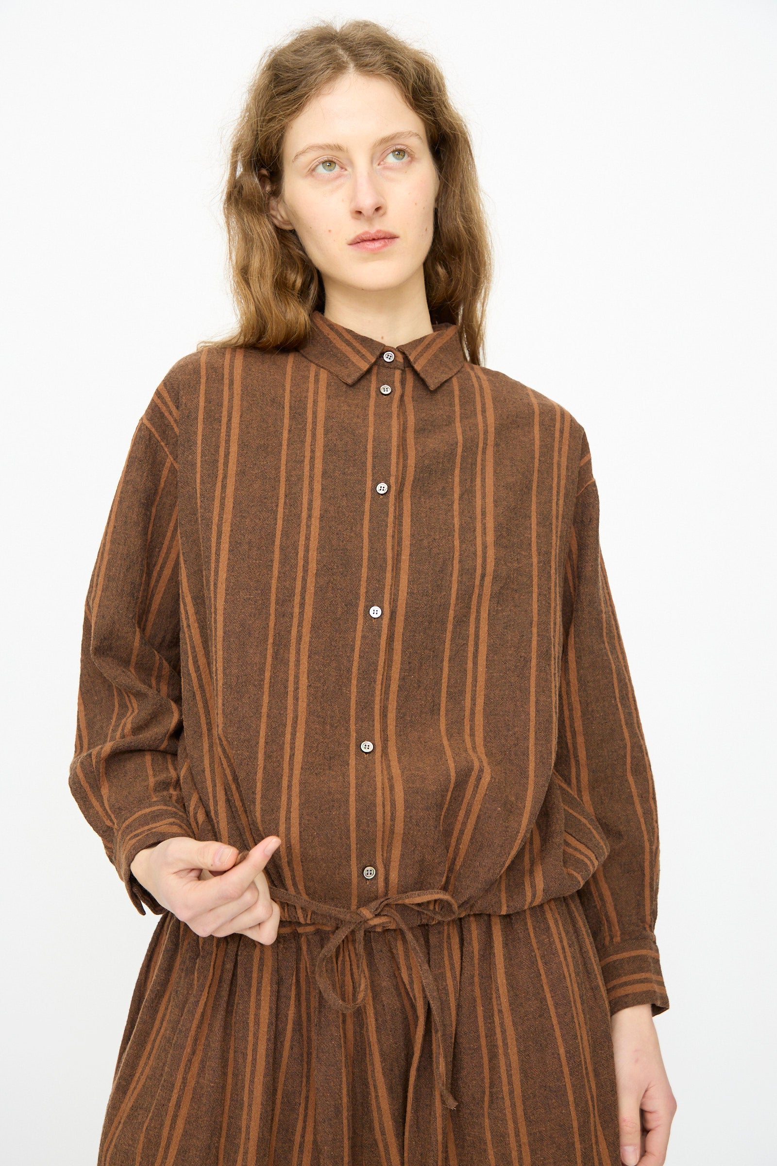 A person with long light brown hair wears the Ichi Antiquités Linen Cotton Random Stripe Shirt in Brown, styled with a waist tie, standing against a plain white background.