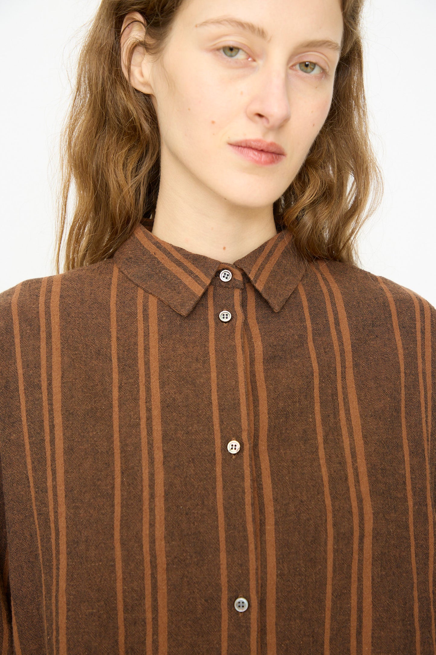 A person with long light brown hair, wearing the Ichi Antiquités Linen Cotton Random Stripe Shirt in Brown—a relaxed-fit button-up with vertical stripes—looks directly at the camera.