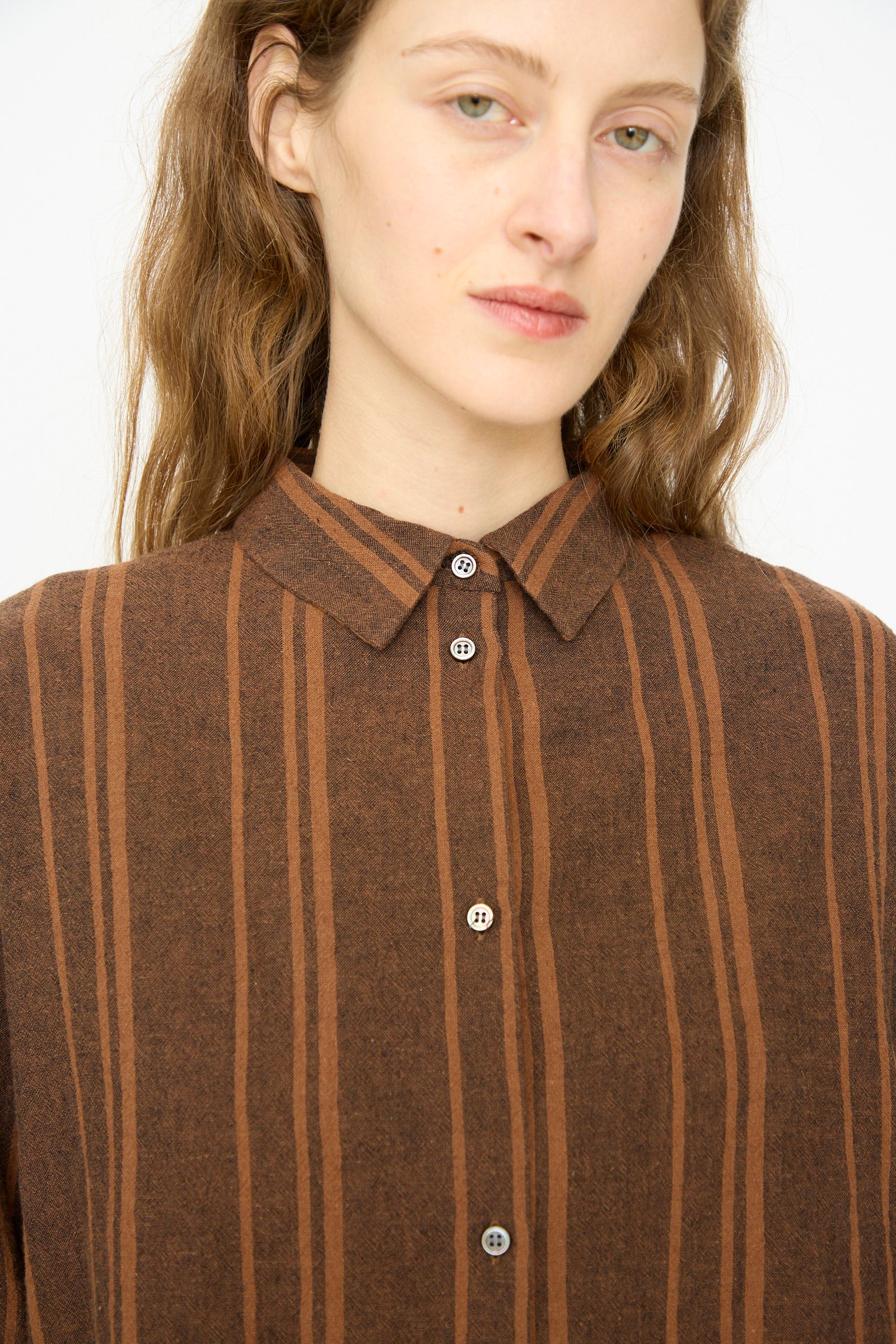 A person with long light brown hair, wearing the Ichi Antiquités Linen Cotton Random Stripe Shirt in Brown—a relaxed-fit button-up with vertical stripes—looks directly at the camera.