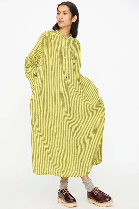 A person wears the Ichi Antiquités Linen Cotton Stripe Dress in Yellow, featuring green vertical stripes, styled with brown shoes and beige socks, standing against a plain white background.